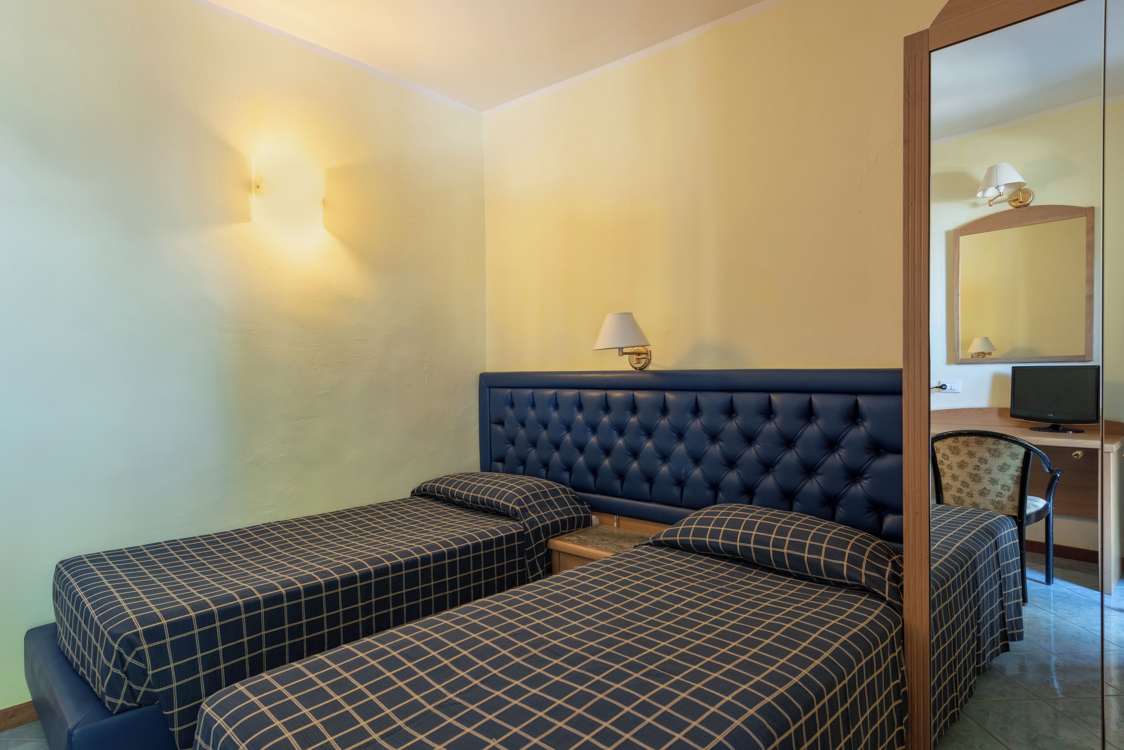 A room with two single beds with checkered bedding, a tufted headboard, a desk with a computer, a chair, and a mirror.