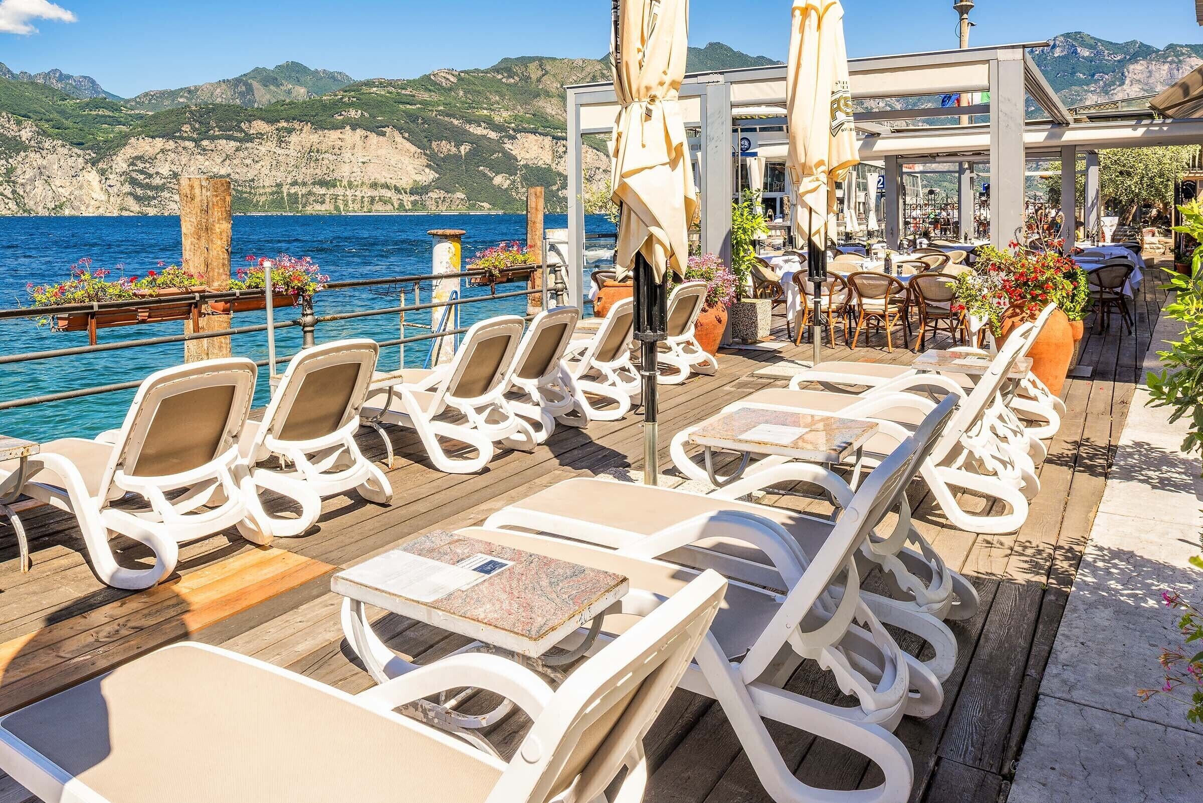 A seaside terrace with sun loungers, tables, and umbrellas, overlooking the sea and mountains.