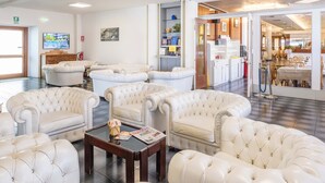 A hotel lobby with plush white leather sofas arranged in a semi-circle, a coffee table with a magazine, and a television screen displaying a hotel image. - Hotel Malcesine (Malcesine)