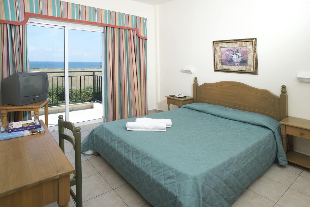 A hotel room with a bed, a television on a wooden table, a nightstand, and a view of the ocean through a sliding door.