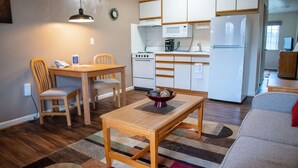 A compact kitchen with white cabinets, a microwave, a stove, and a refrigerator. There is a dining table with chairs and a small rug on the floor.