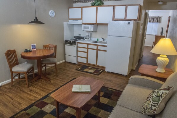 Un espace de vie compact comprenant un coin cuisine, un coin repas avec une table et des chaises, un canapé avec un coussin et une lampe sur une table d’appoint.