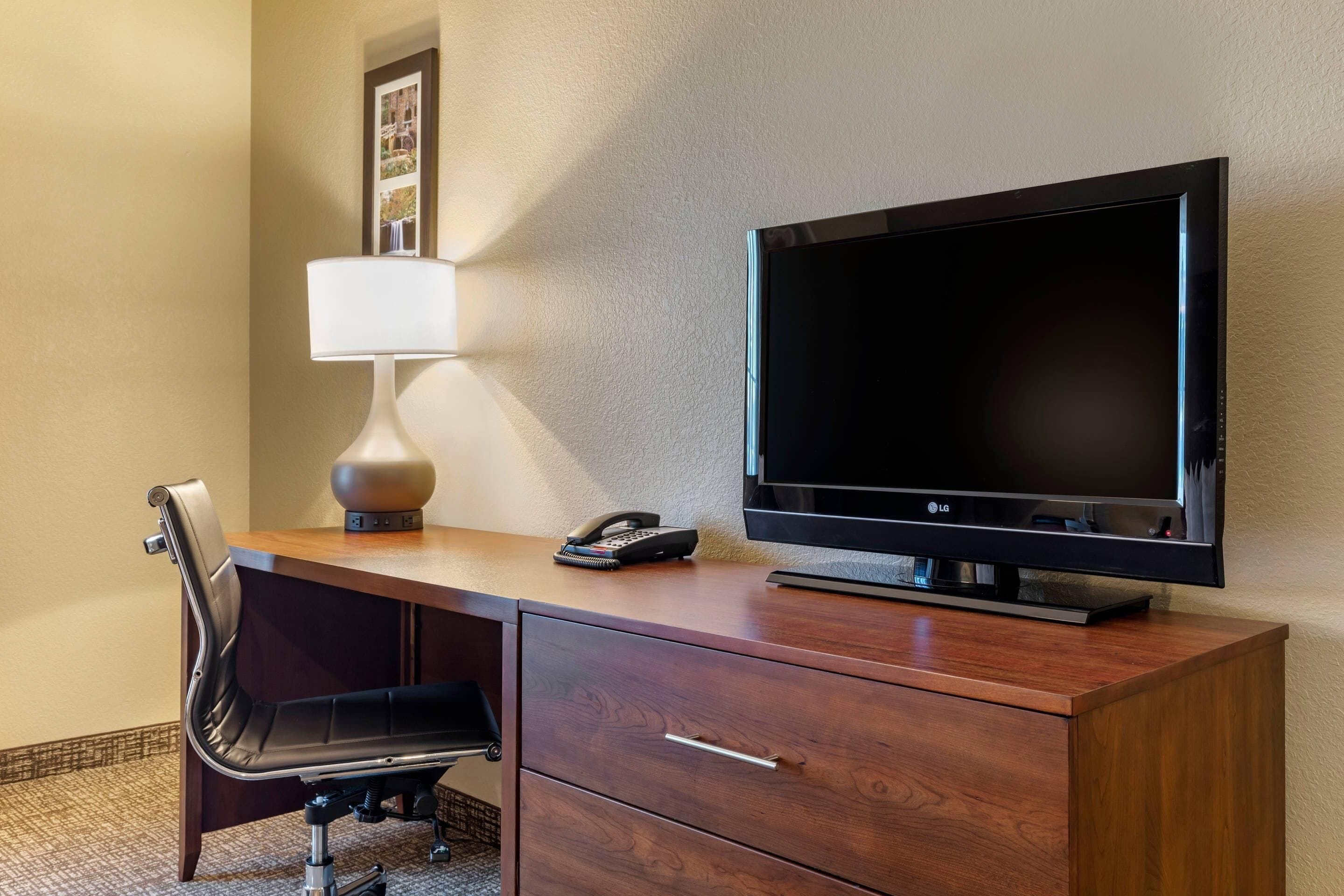 A hotel room with a wooden desk, a black office chair, a flat-screen TV, a lamp, and a telephone.