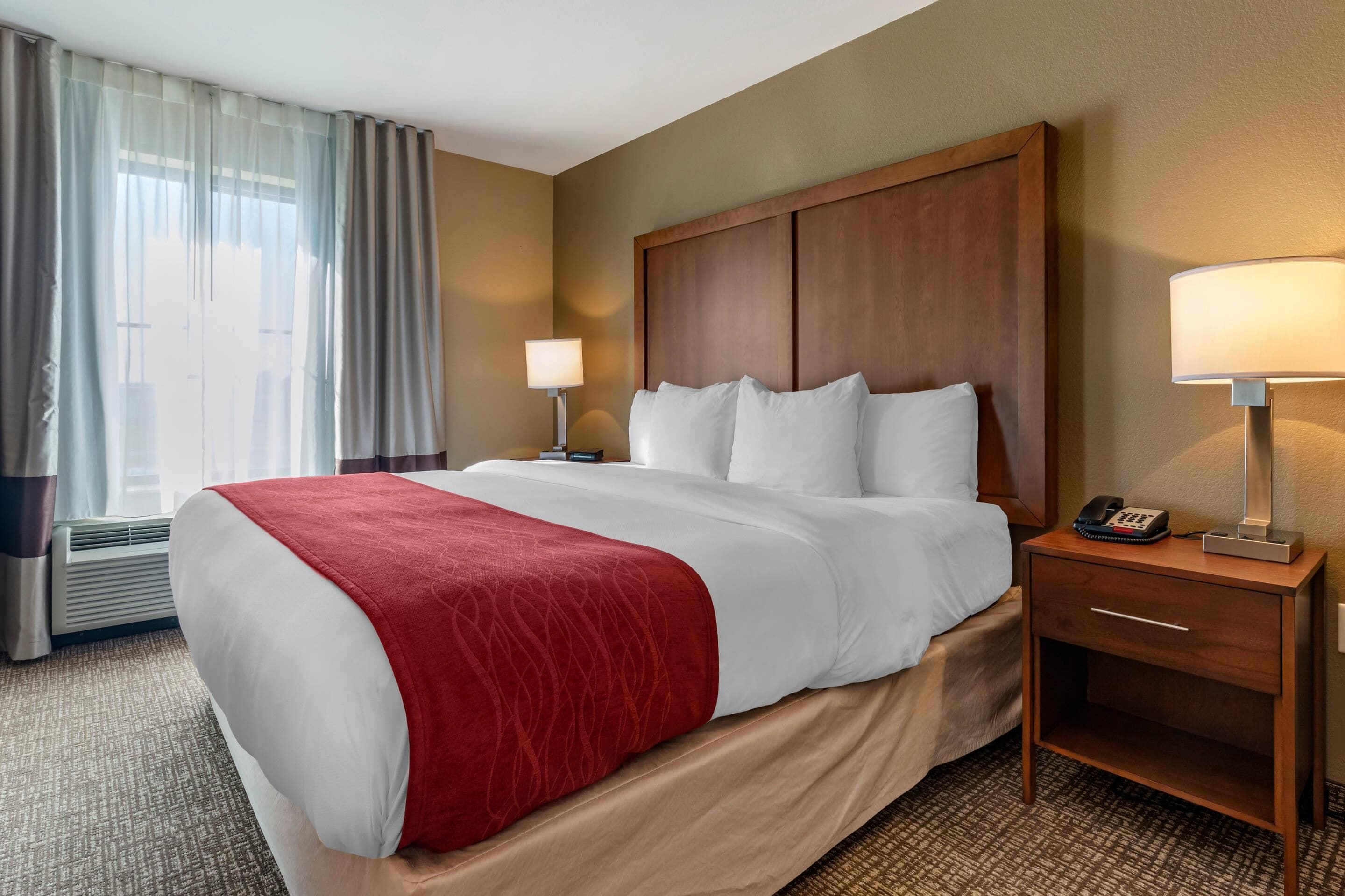 A hotel room with a large bed, a bedside table with a lamp, a telephone, and a window with curtains.