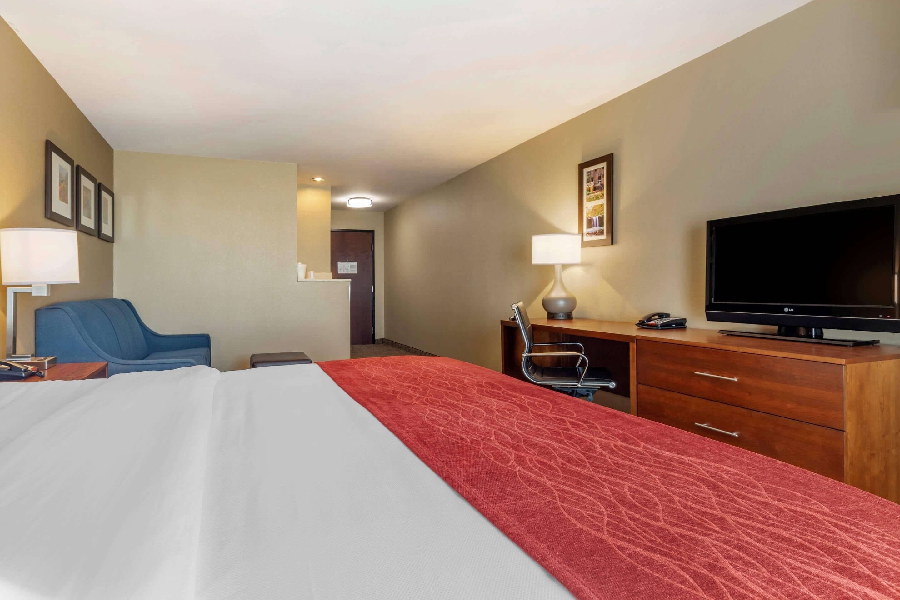 A hotel room with a bed, a desk with a television, a chair, and a desk lamp.