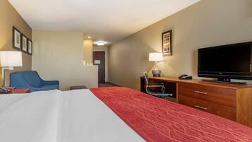 A hotel room with a bed, a desk with a television, a chair, and a desk lamp.