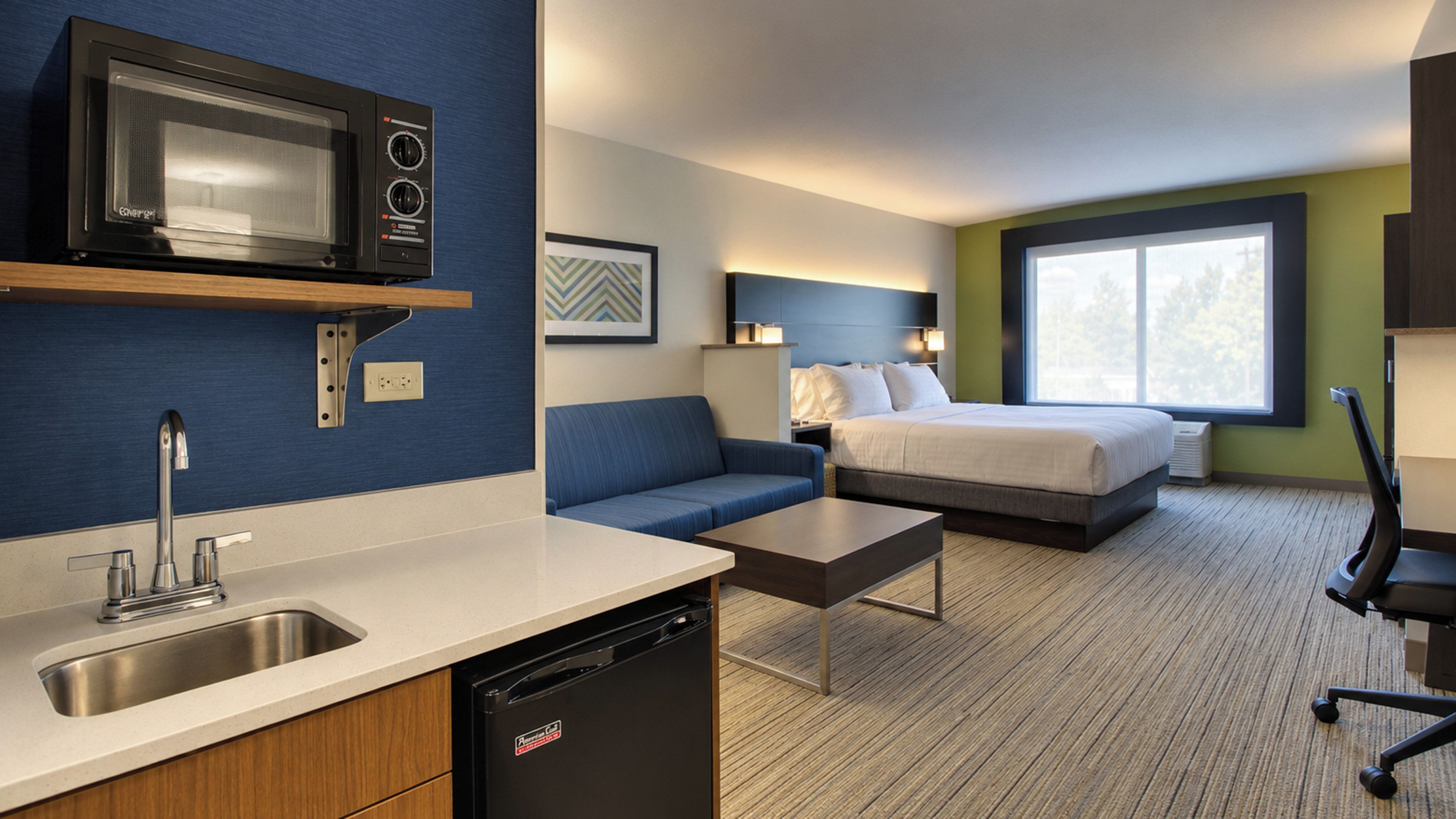 A modern hotel room with a kitchenette, a bed with white linens, a blue sofa, and a large window providing natural light.