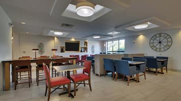 A modern dining area with multiple tables and chairs, recessed ceiling lights, and a large wall clock.