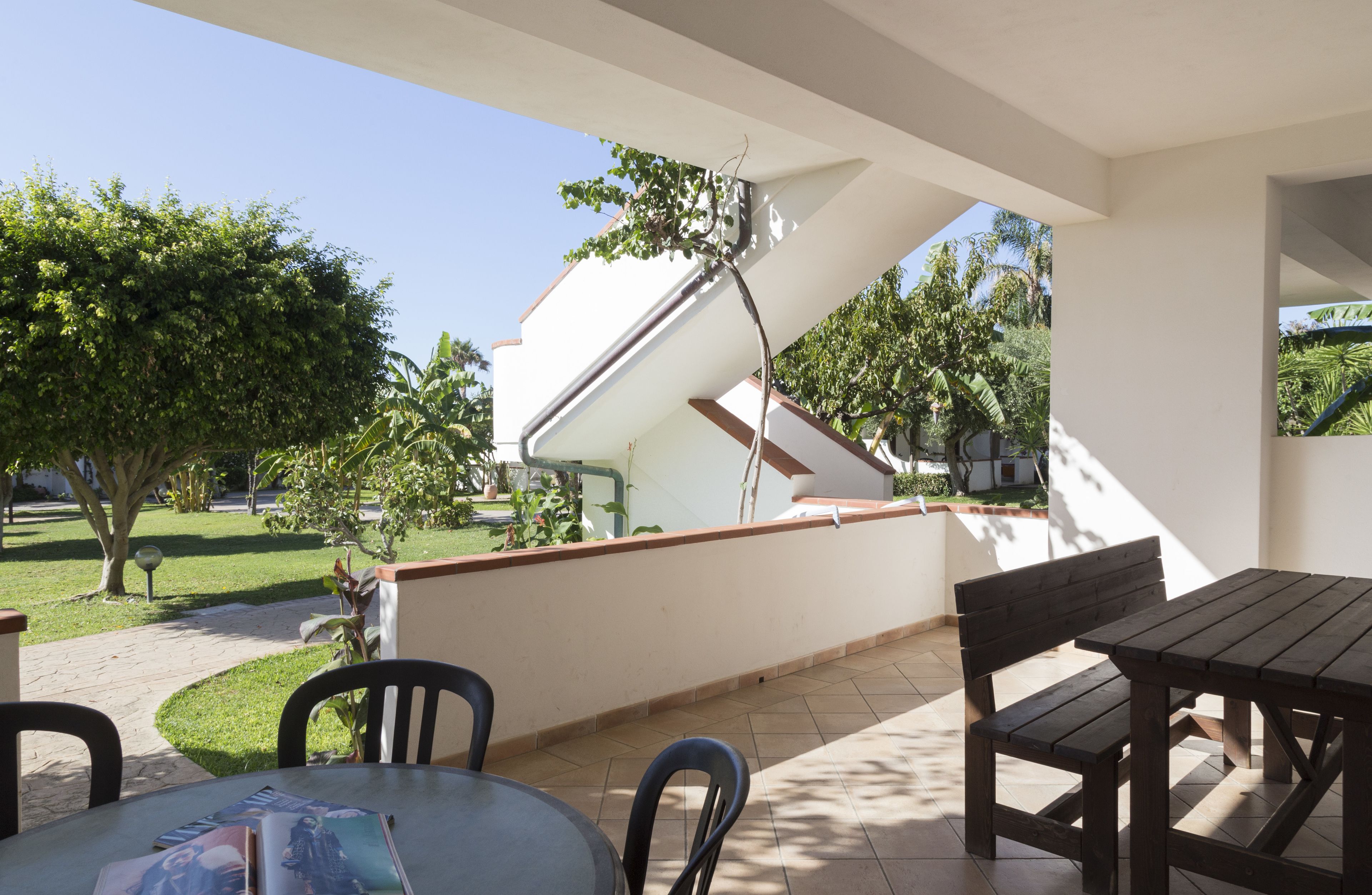 An outdoor patio area with a table and chairs, a covered section with a white wall and a red roof, and a view of a garden and trees.