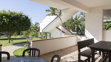 An outdoor patio area with a table and chairs, a covered section with a white wall and a red roof, and a view of a garden and trees.