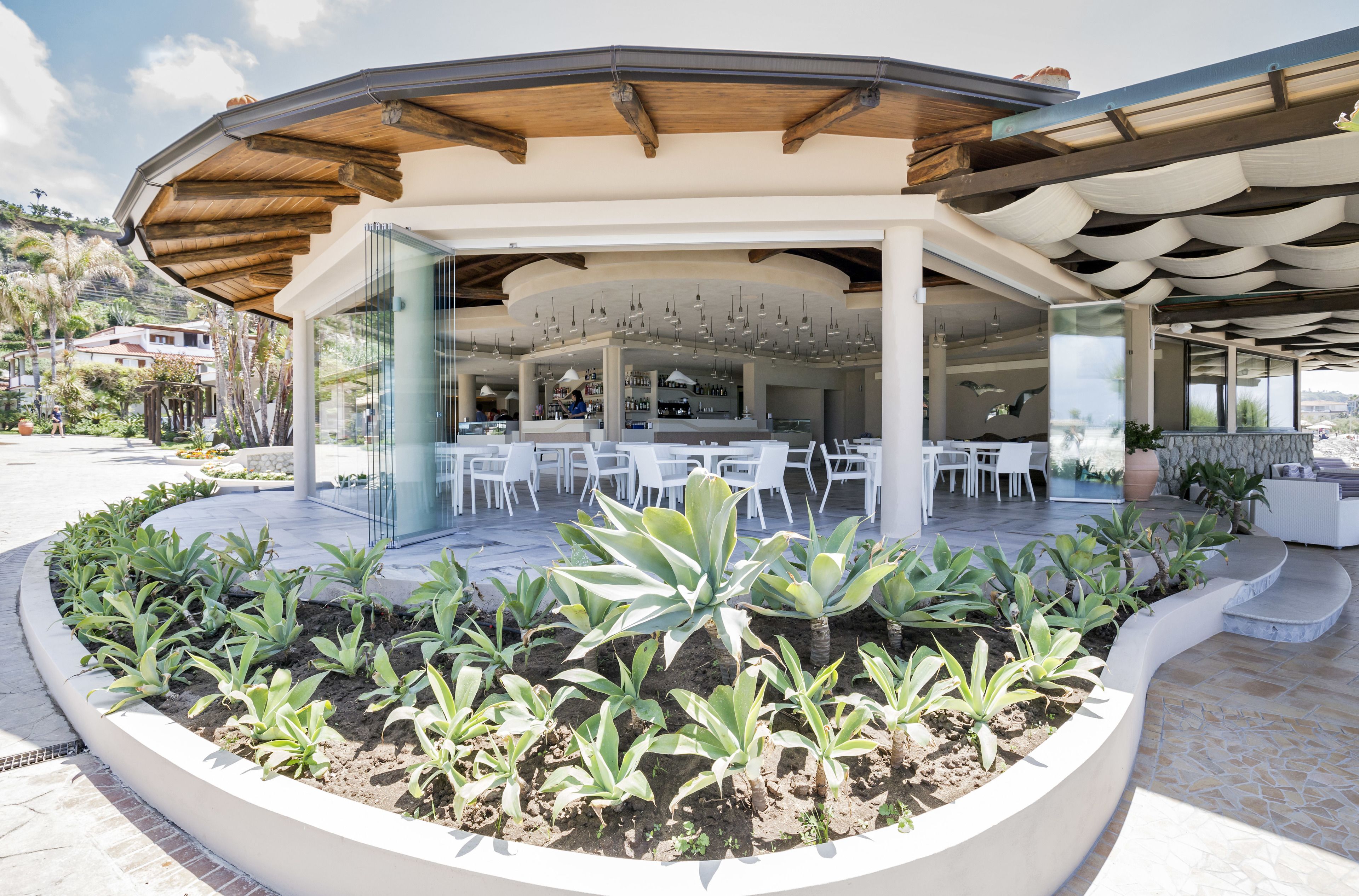 Un espace extérieur aménagé, abrité et doté de tables et de chaises blanches, entouré d’un jardin paysager avec différentes espèces de plantes.