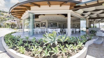 Un espace extérieur aménagé, abrité et doté de tables et de chaises blanches, entouré d’un jardin paysager avec différentes espèces de plantes.