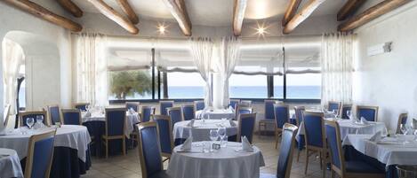 Un restaurant doté de nombreuses tables et chaises, de grandes fenêtres avec des rideaux et offrant une vue sur la mer.