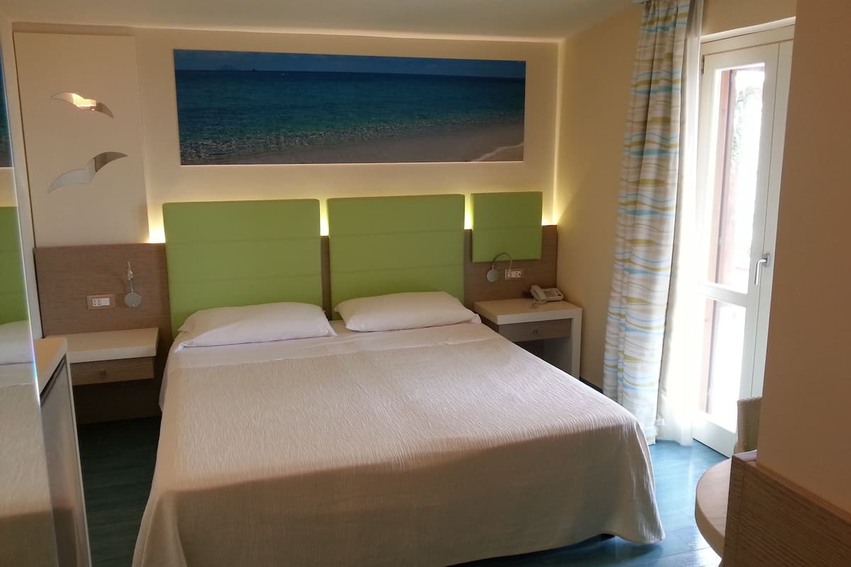 A hotel room with a large bed, two bedside tables, a nightstand, a painting of a beach, and a view of the sea through the window.