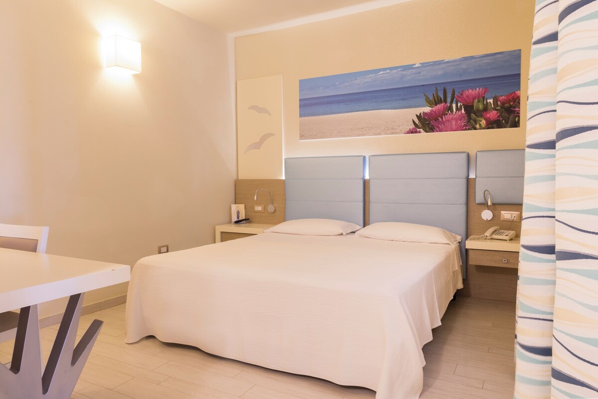 A hotel room with a bed, bedside tables, a desk, and a painting of a beach scene.