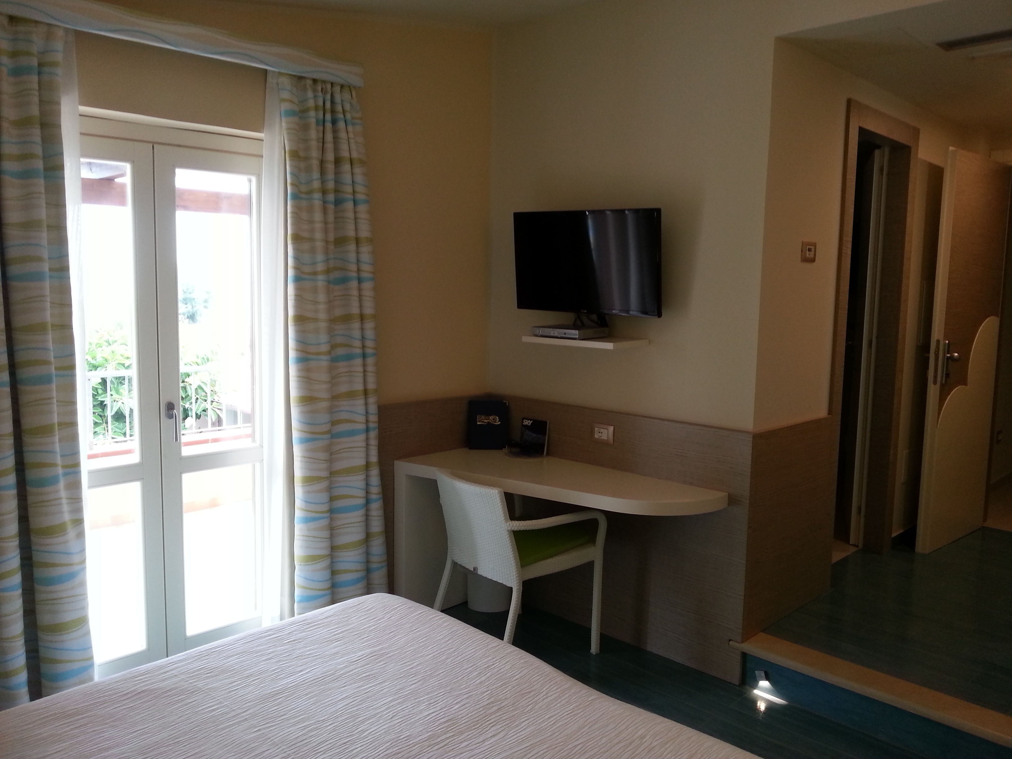 A hotel room with a bed, a desk with a chair, a television mounted on the wall, and a sliding door leading to an outdoor area.