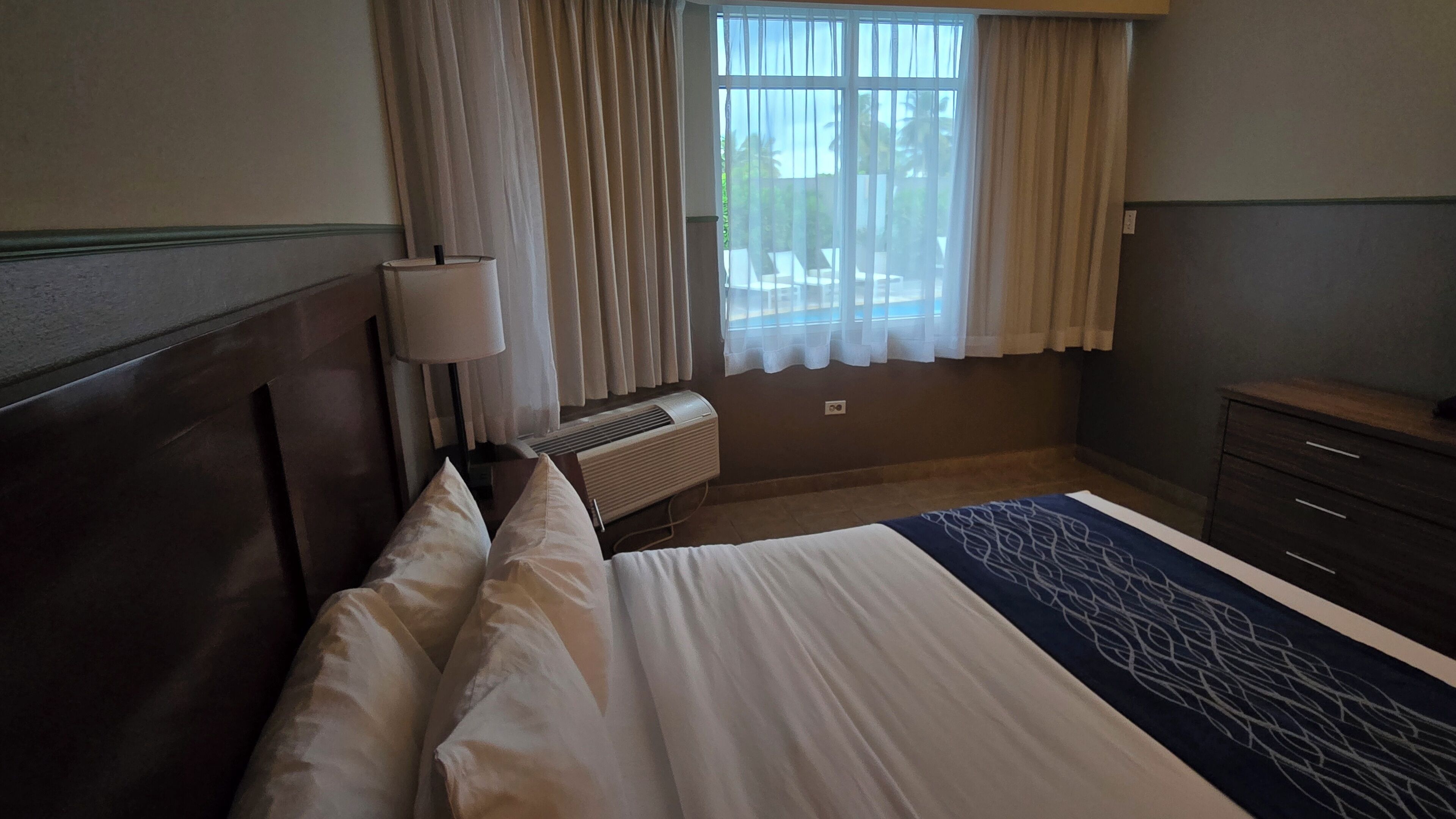 A hotel room with a bed, a bedside lamp, a window with curtains, an air conditioning unit, and a dresser.