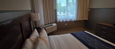 Une chambre d’hôtel comprenant un lit, une lampe de chevet, une fenêtre avec des rideaux, un appareil de climatisation et une commode.