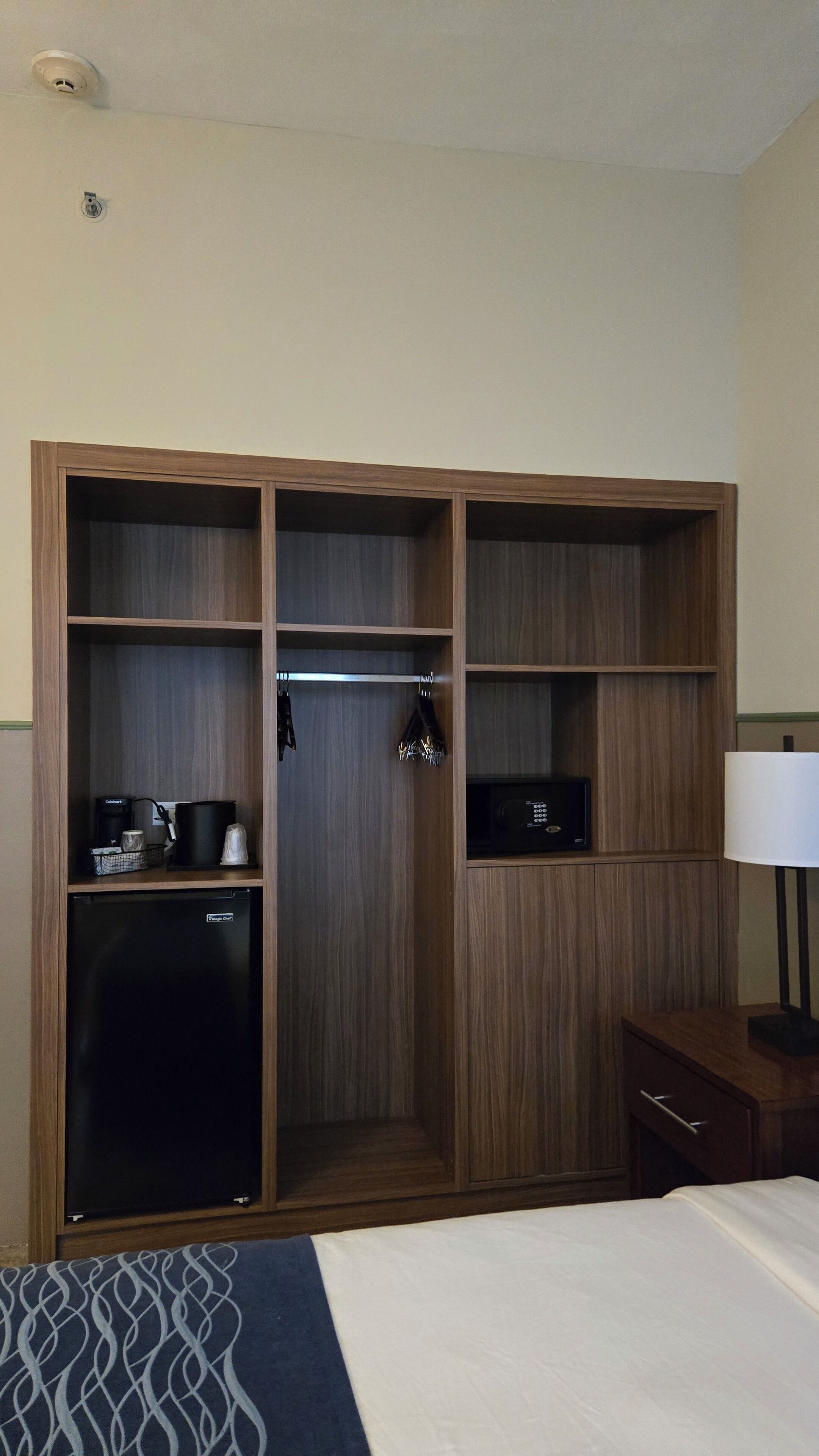 Habitación de hotel con un armario grande, una cama, un escritorio con lámpara y una cafetera.