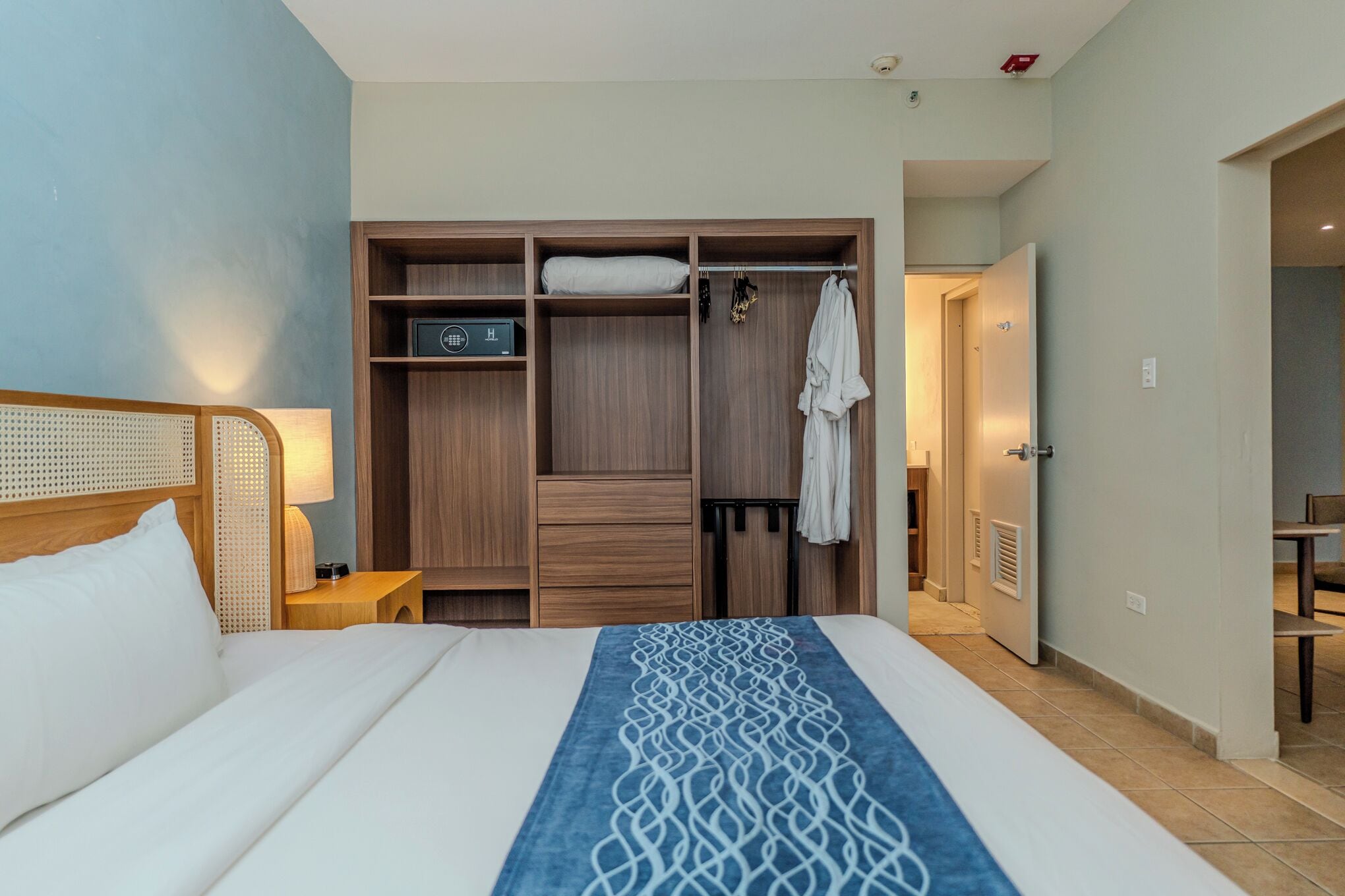 Habitación de hotel con cama, armario de madera, mesita de noche y baño visible a través de una puerta abierta.
