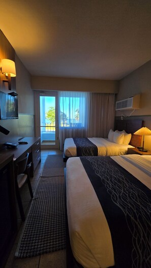 Habitación de hotel con dos camas, un escritorio, televisión y balcón con vistas.