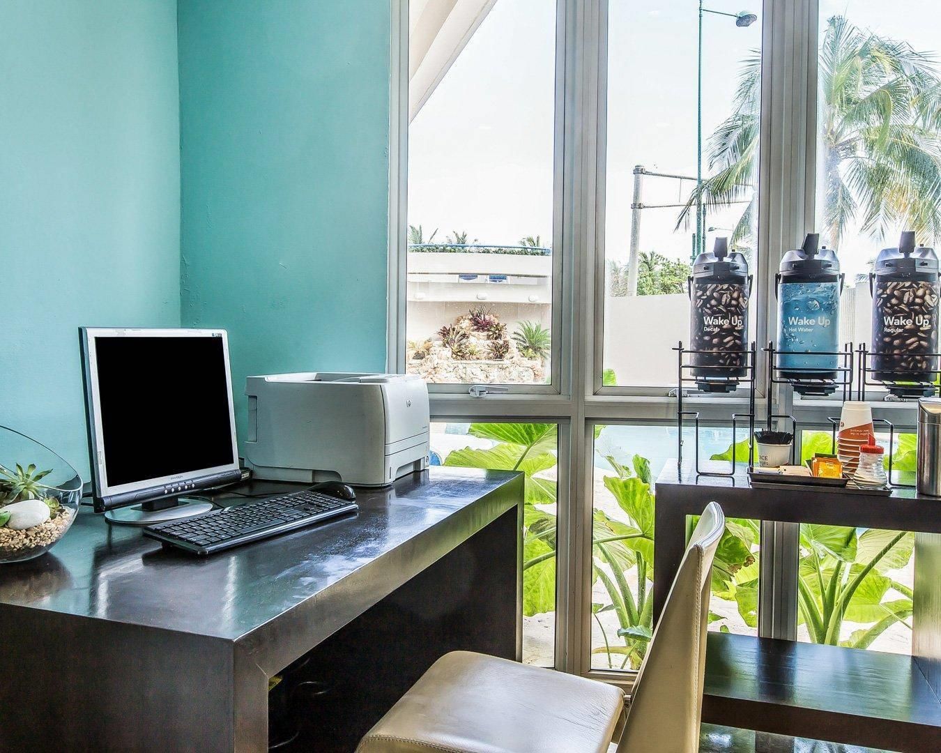 Un espacio de trabajo con computadora de escritorio, impresora y vista a una piscina y palmeras a través de amplias ventanas.