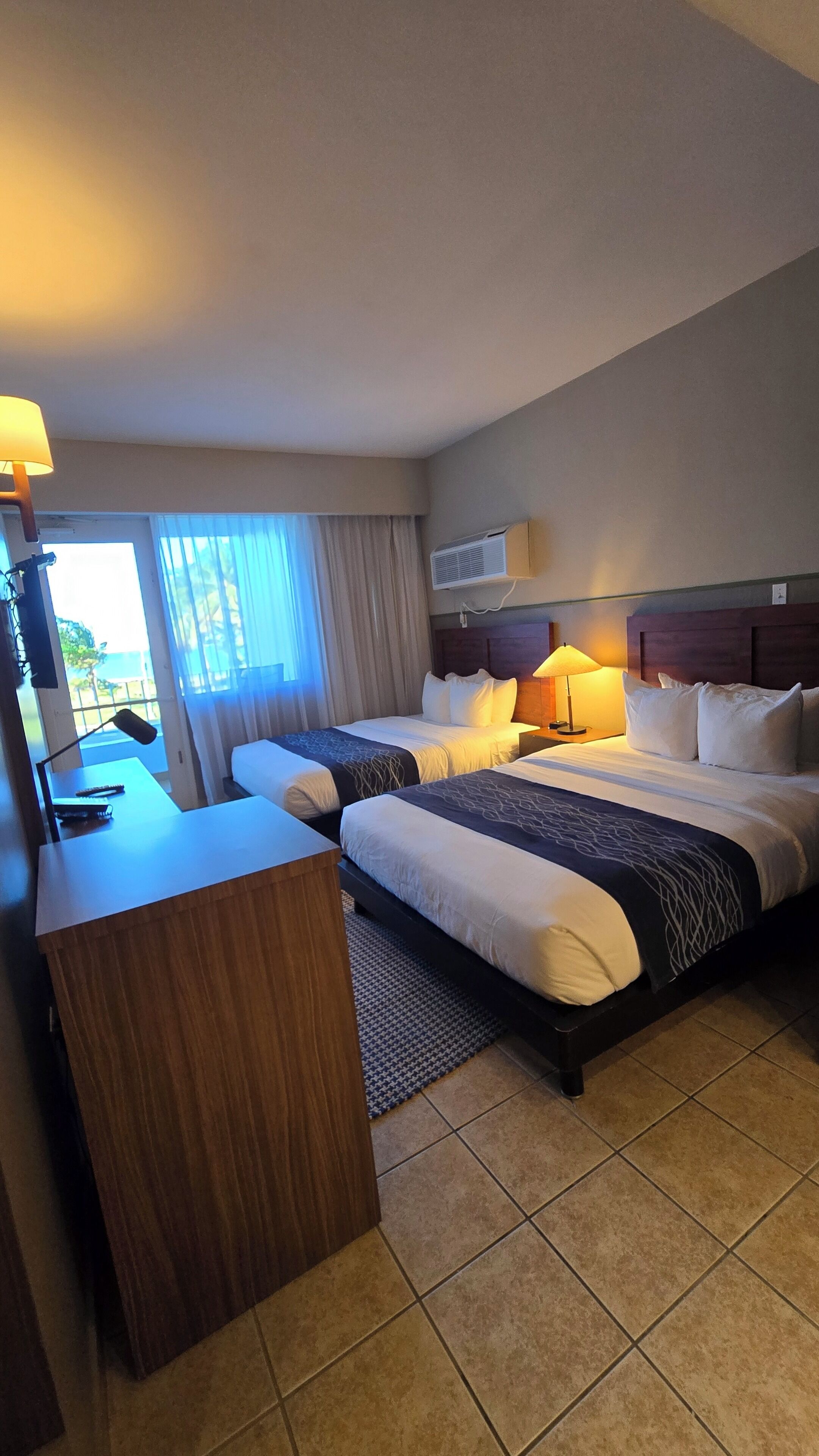 Une chambre d’hôtel avec deux lits, un bureau, une lampe, un appareil de climatisation et une vue sur l’extérieur.