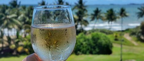 Una mano sosteniendo una copa de vino blanco, con vista a una playa y palmeras al fondo.