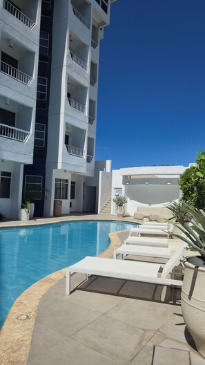 Un edificio moderno con área de piscina, que incluye sillas de descanso y plantas en macetas.
