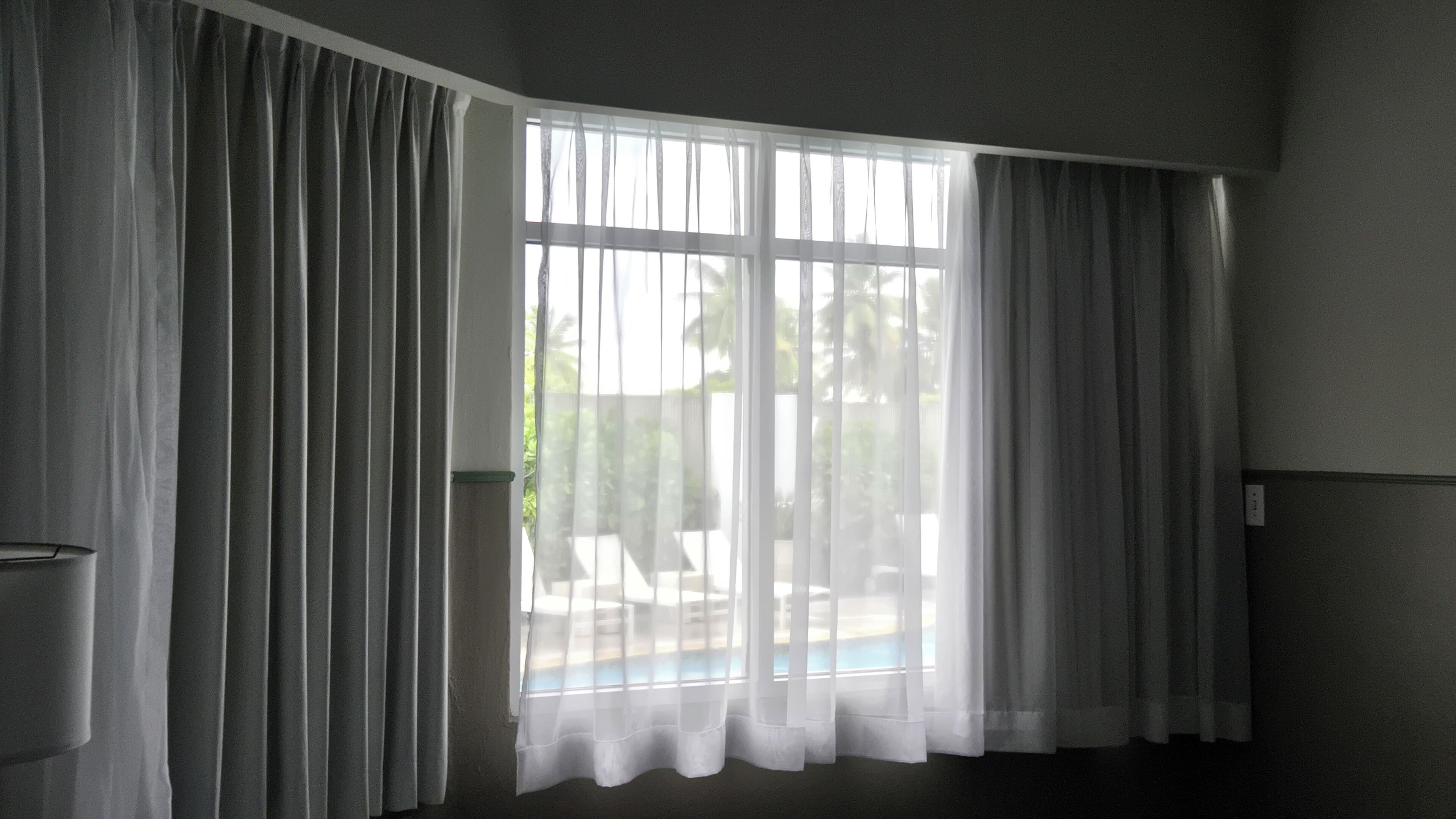 A room with a large window covered by sheer curtains, a dark floor, and a visible outdoor area with a pool.