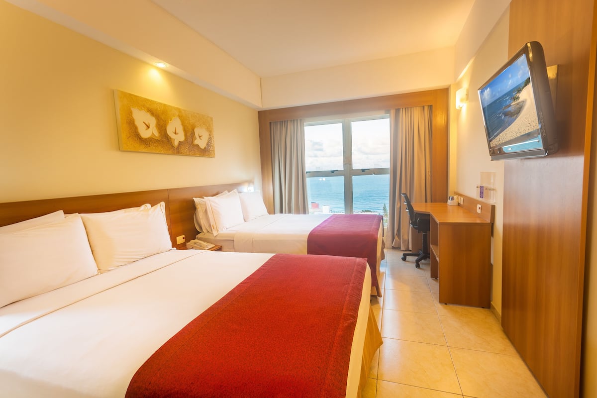 A hotel room with two beds, a desk, a TV, and a large window with a view of the sea.