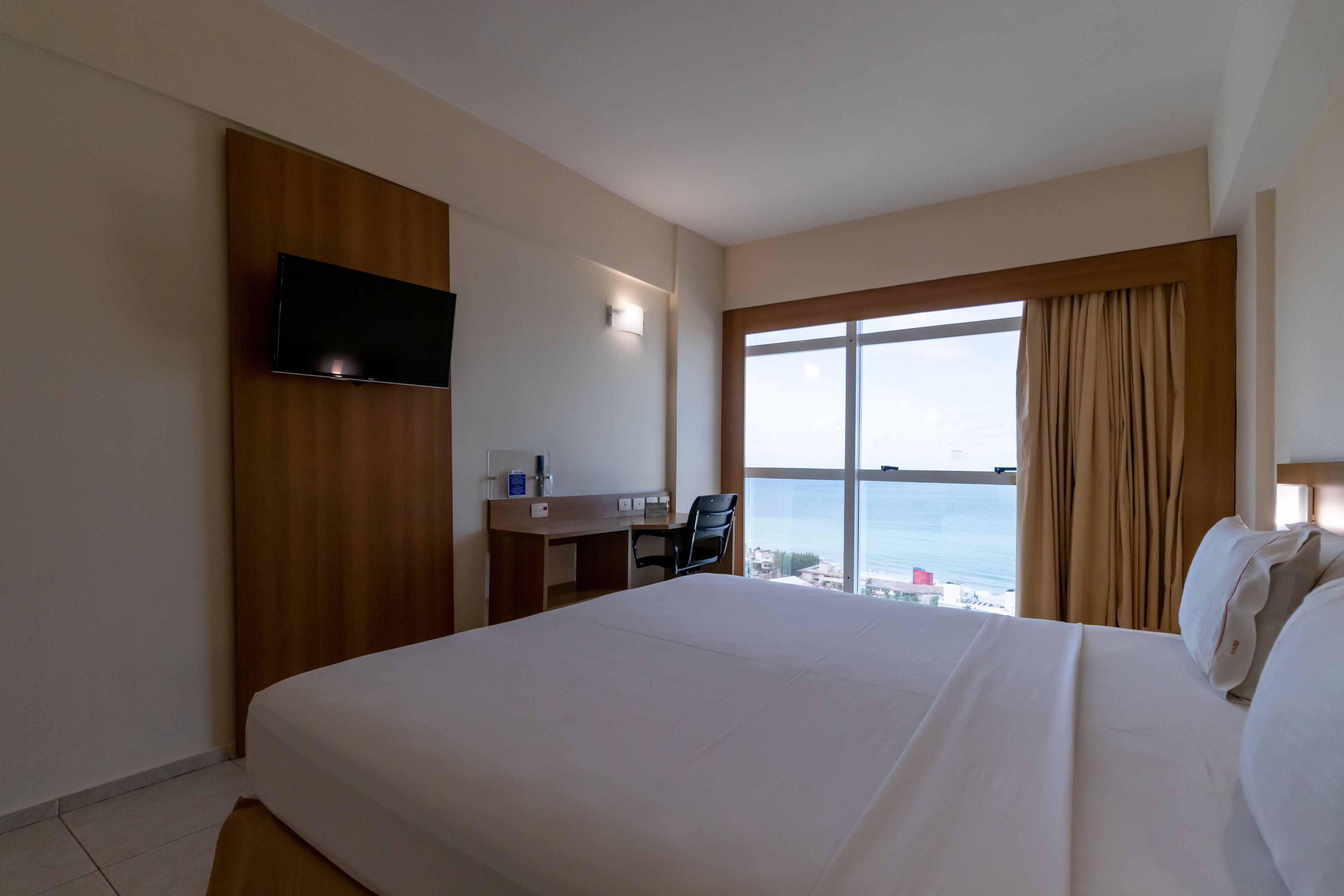 Quarto de hotel com uma cama grande, uma escrivaninha, uma cadeira, uma televisão de tela plana na parede e uma janela com cortinas.