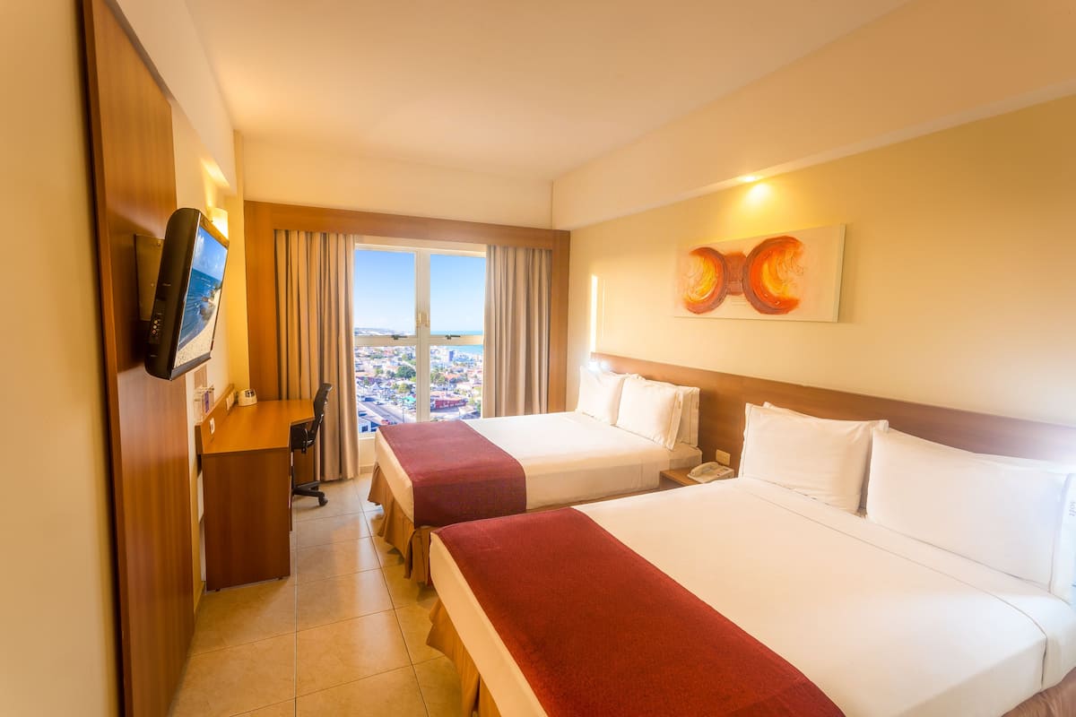 A hotel room with two beds, a desk, a TV, and a large window with a city view.
