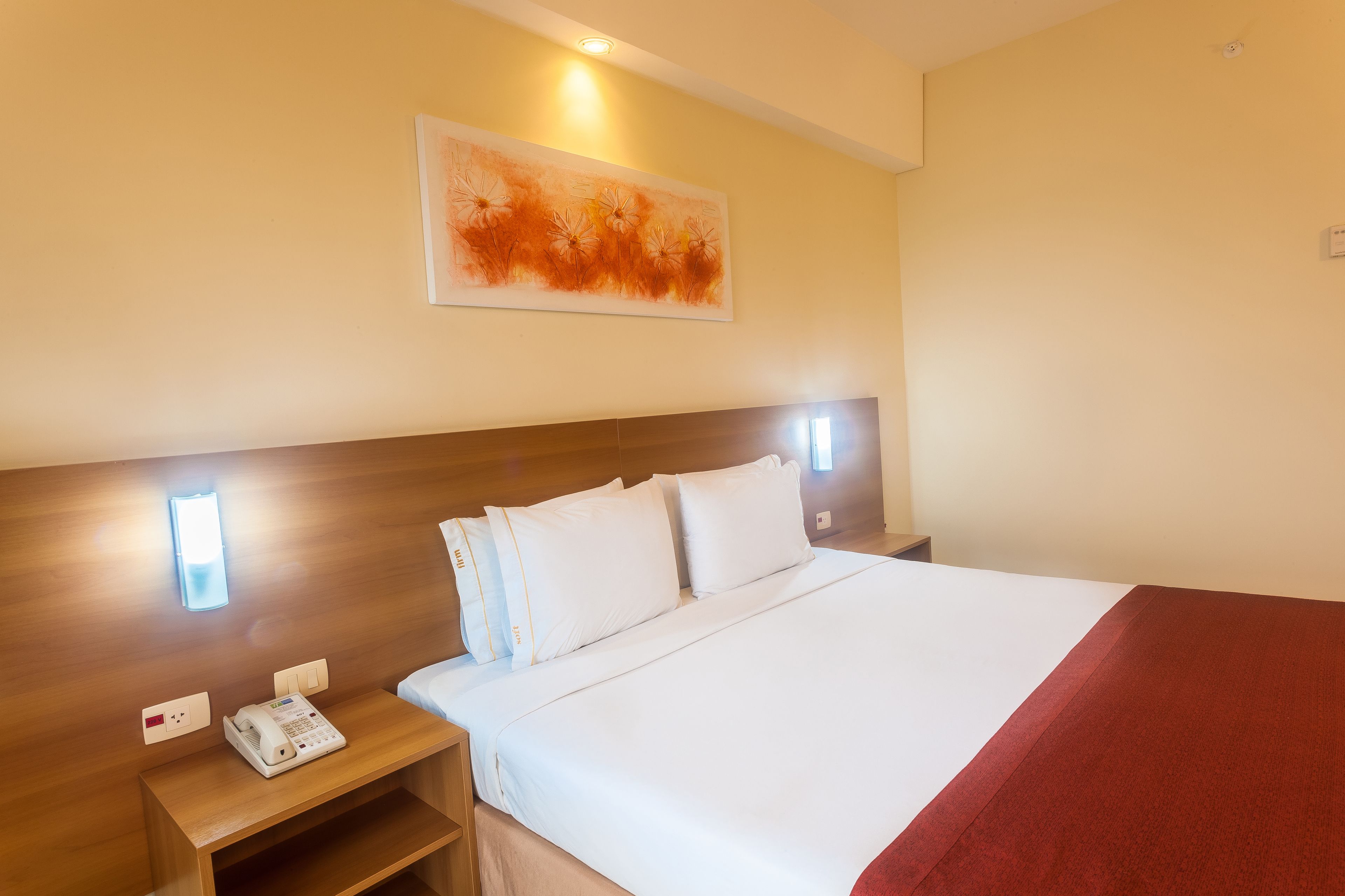 Quarto de hotel com cama bem arrumada, mesa de cabeceira com telefone e luminária na parede.
