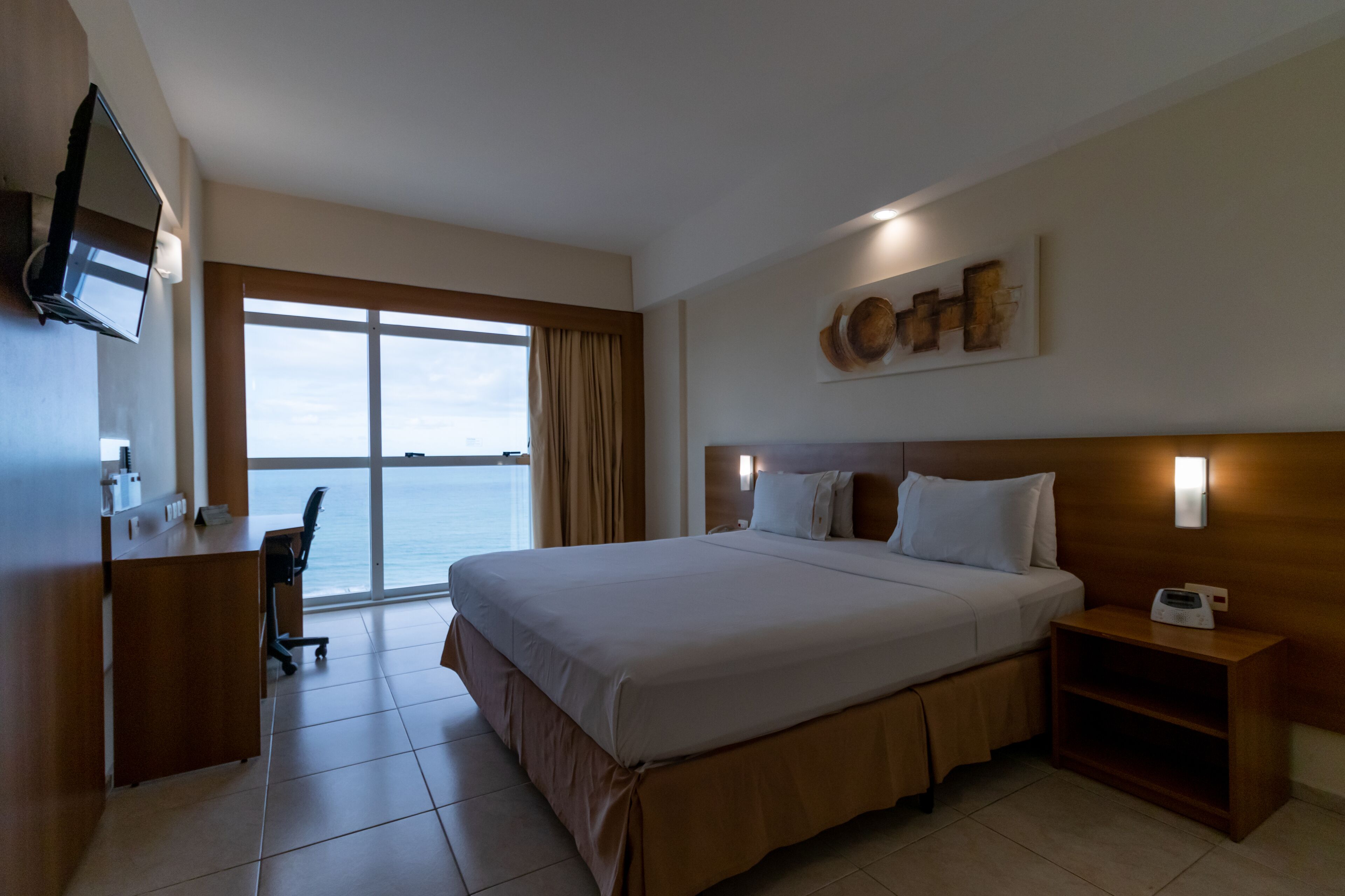 A hotel room with a large bed, a desk with a chair, a television, and a view of the sea through a large window.