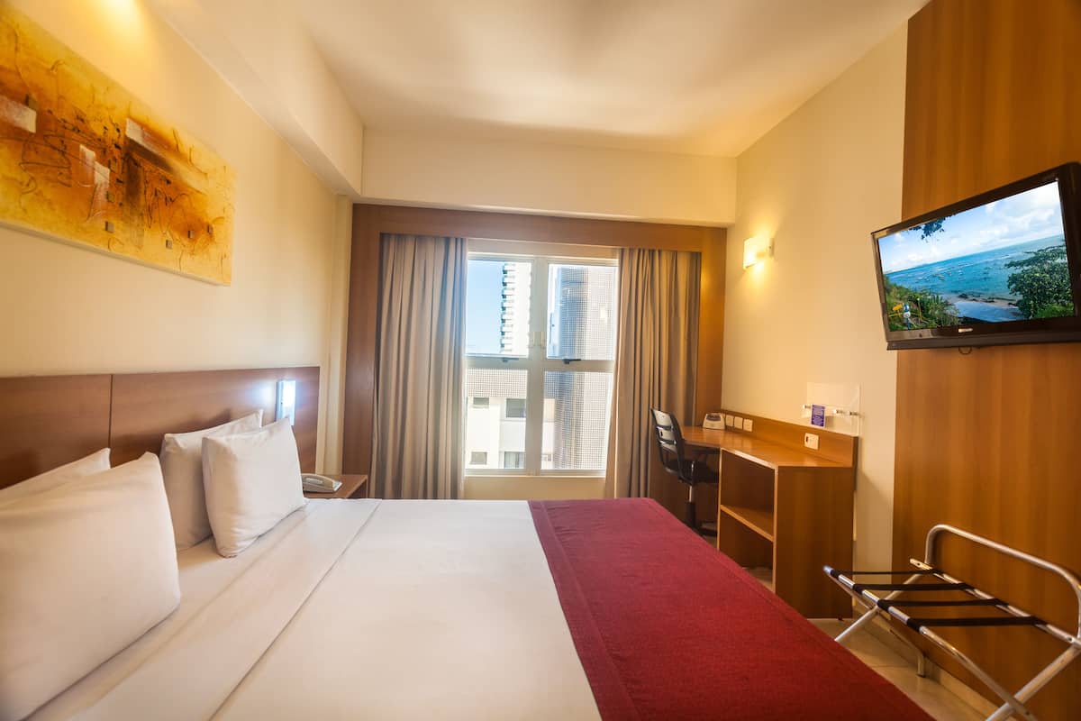 A hotel room with a large bed, a desk, a television, and a view of the city through the window.