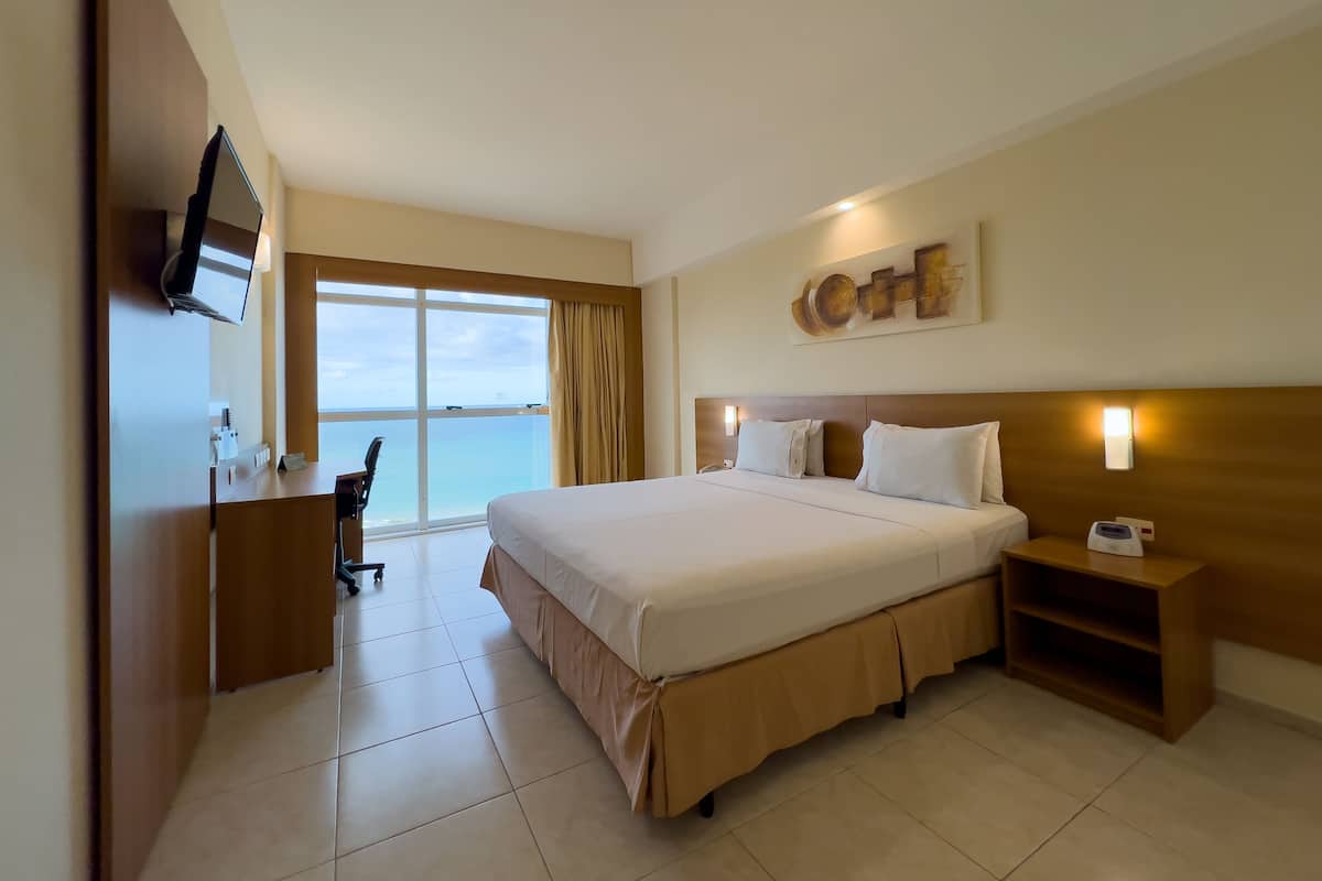 A hotel room with a large bed, a desk with a chair, a TV mounted on the wall, and a view of the ocean through a large window.