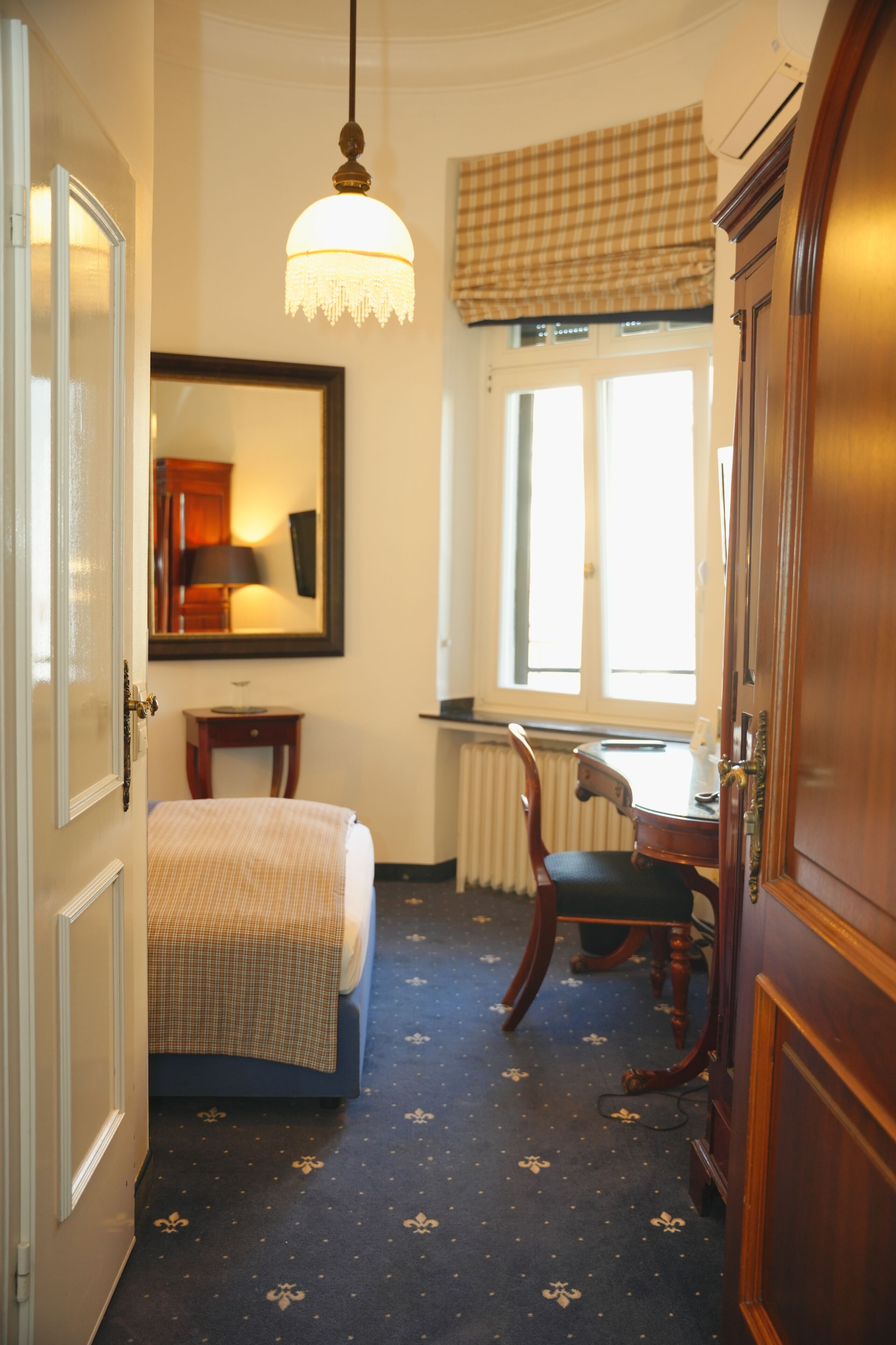 A narrow hotel room with a bed, a desk, a chair, a mirror, and a window with a checkered blind.