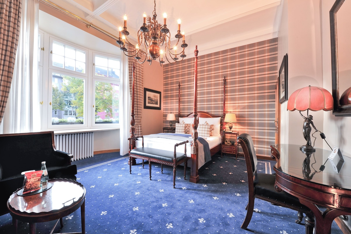 Una habitación con una cama grande, una araña, vistas por la ventana y una alfombra azul.