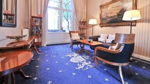 A room with a blue carpet featuring a white coat of arms, a wooden coffee table, a sofa, a chair, a bookshelf, and a painting on the wall. - Hotel Windsor (Düsseldorf)