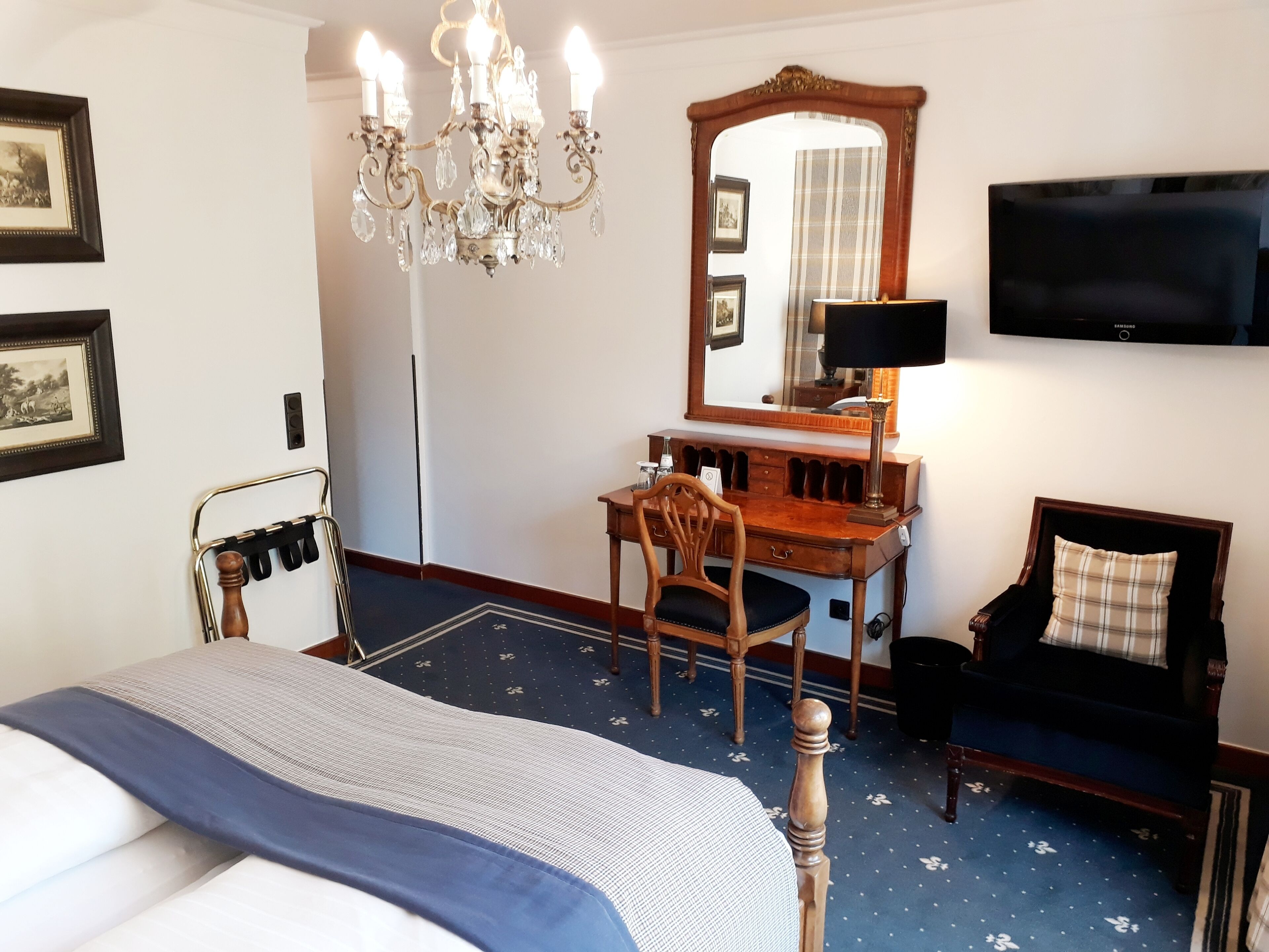 A hotel room with a bed, a desk with a chair, a television, and a chandelier.