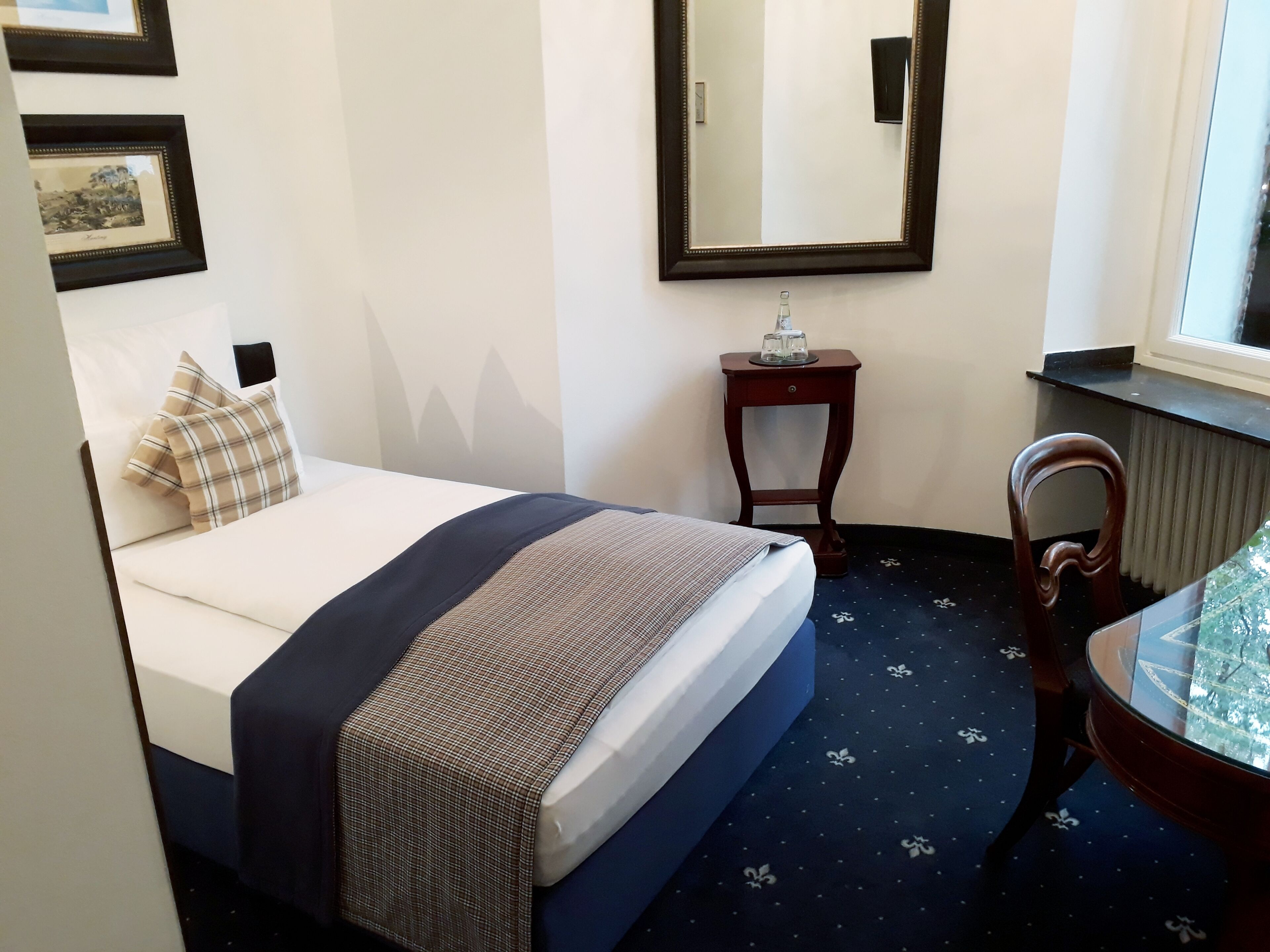 Una habitación de hotel con una cama, una mesita con superficie de vidrio, una silla, un espejo y una ventana.