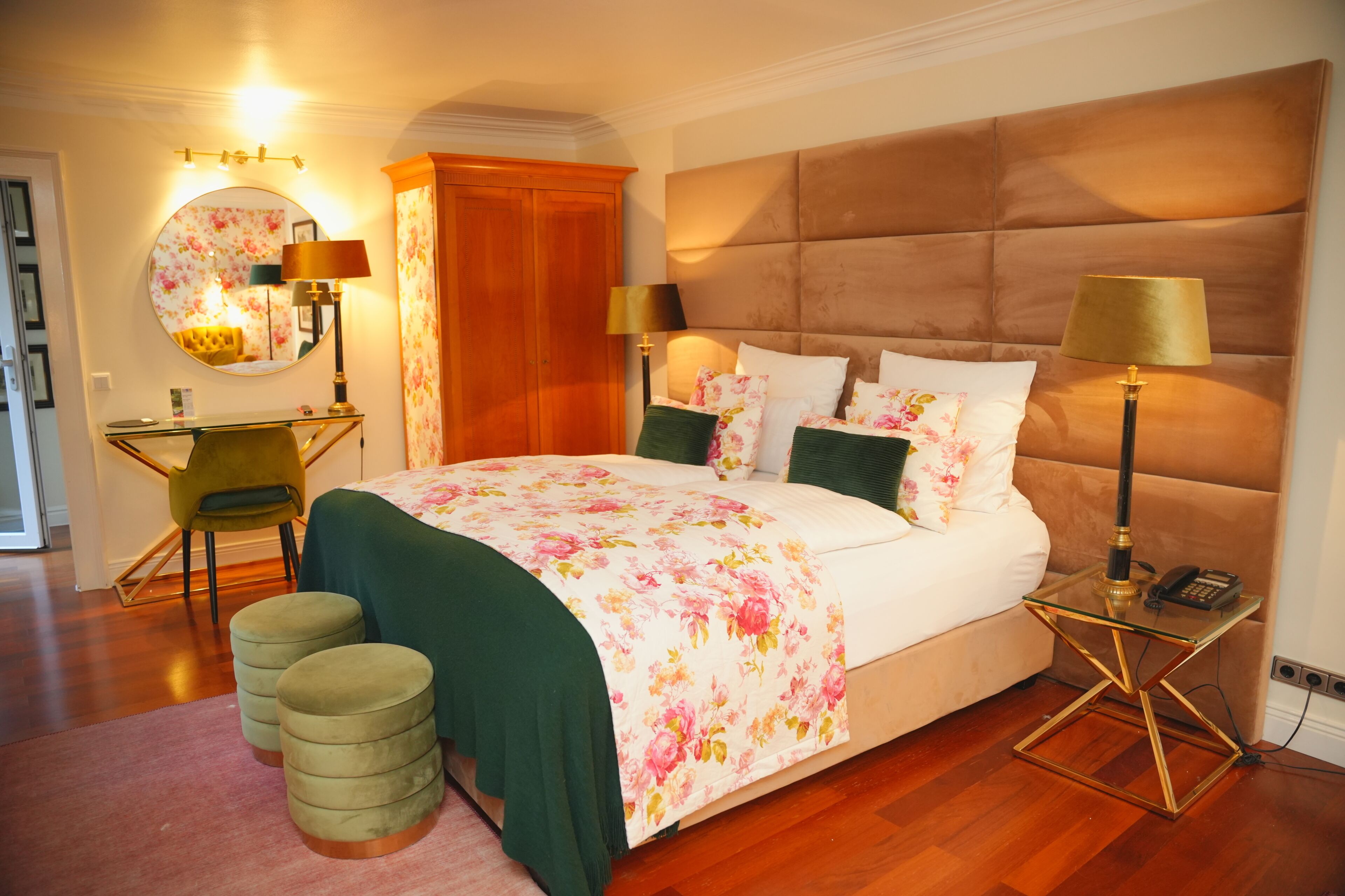Een goed verlichte slaapkamer met een groot bed, twee bedlampen, een bureau met stoel, en een ronde spiegel aan de muur.