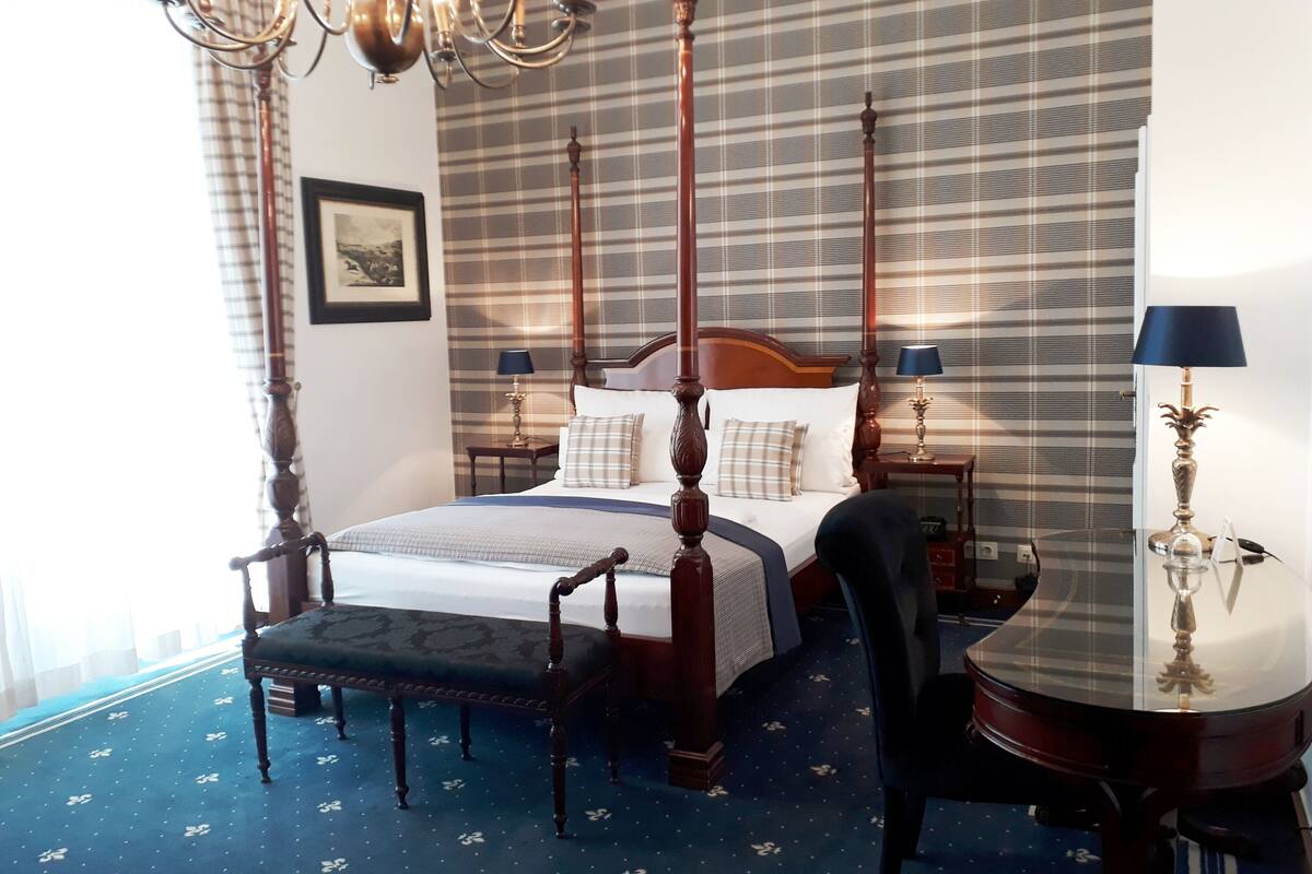 A four-poster bed with a wooden frame, two bedside tables with lamps, a chair, and a patterned carpet.