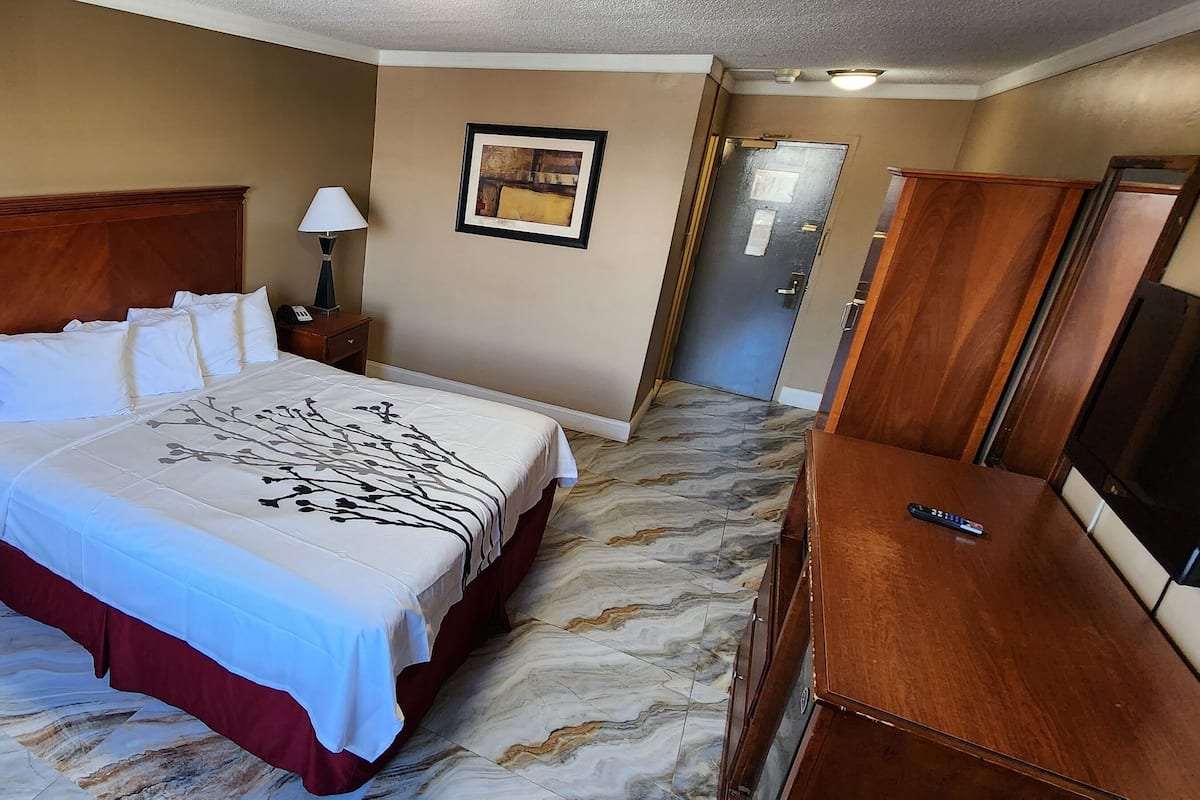 A hotel room with a bed, two bedside tables with lamps, a dresser, a framed picture on the wall, and a door leading to another room.