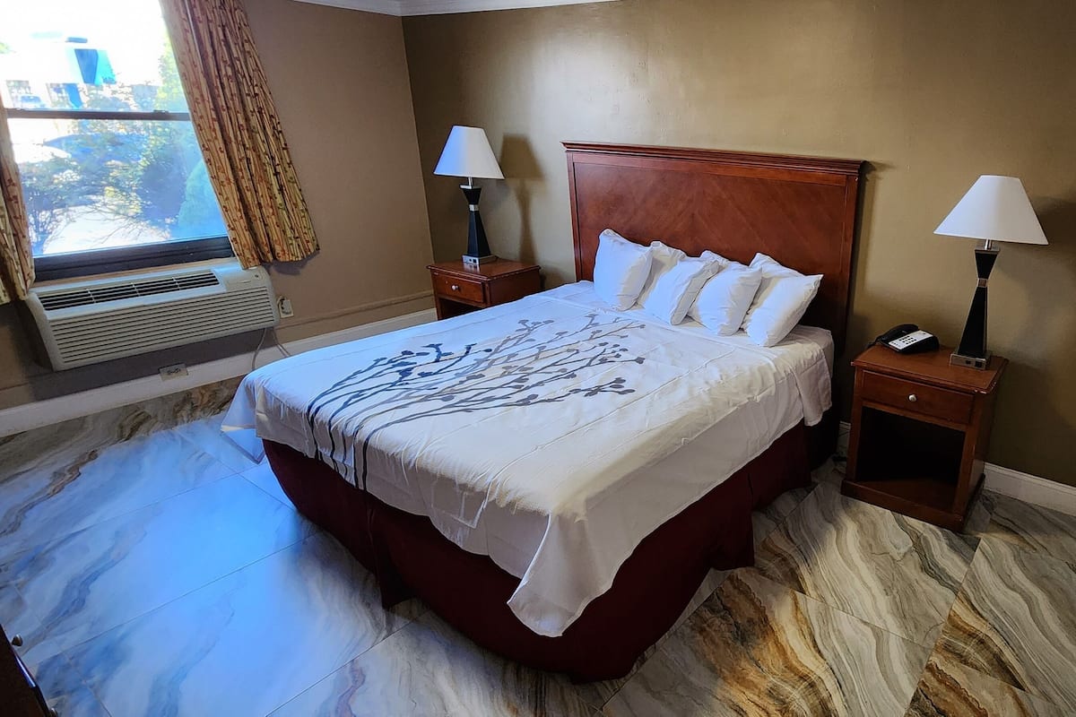 A hotel room with a large bed, two bedside tables with lamps, a wall-mounted air conditioner, and a window with curtains.