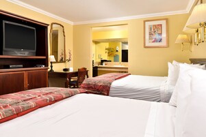 Habitación de hotel con dos camas, televisor, escritorio y baño visible a través de una puerta abierta.