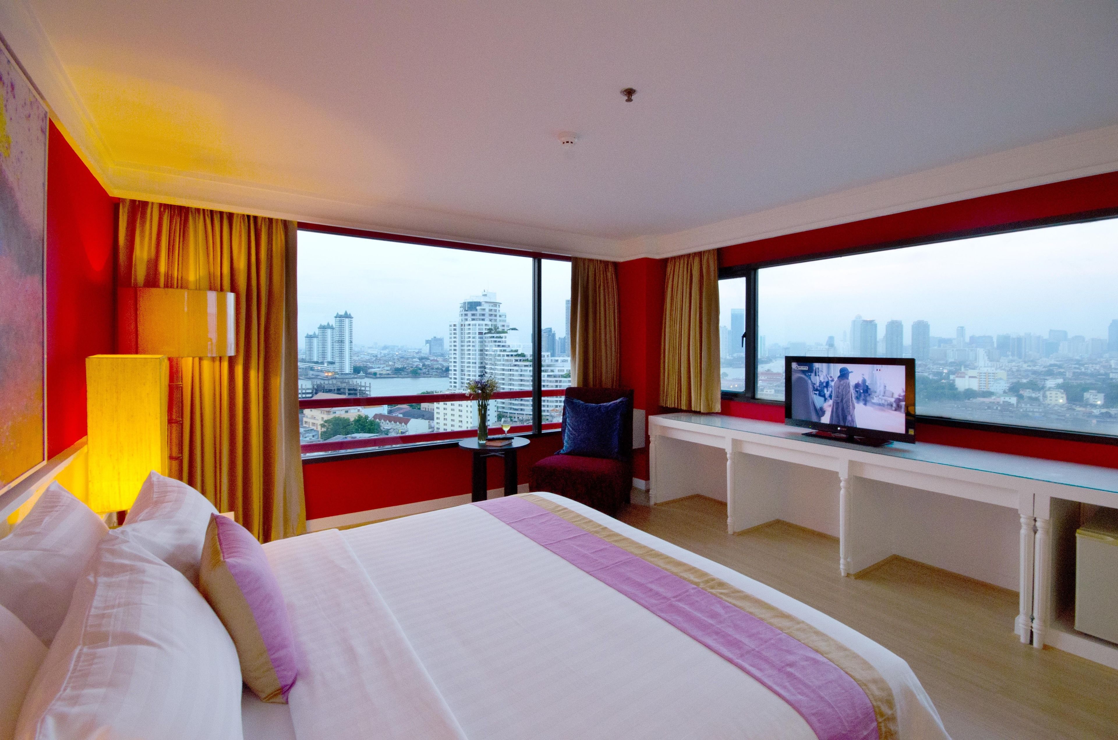 A hotel room with a large bed, a desk, a television, and a view of a cityscape through large windows.