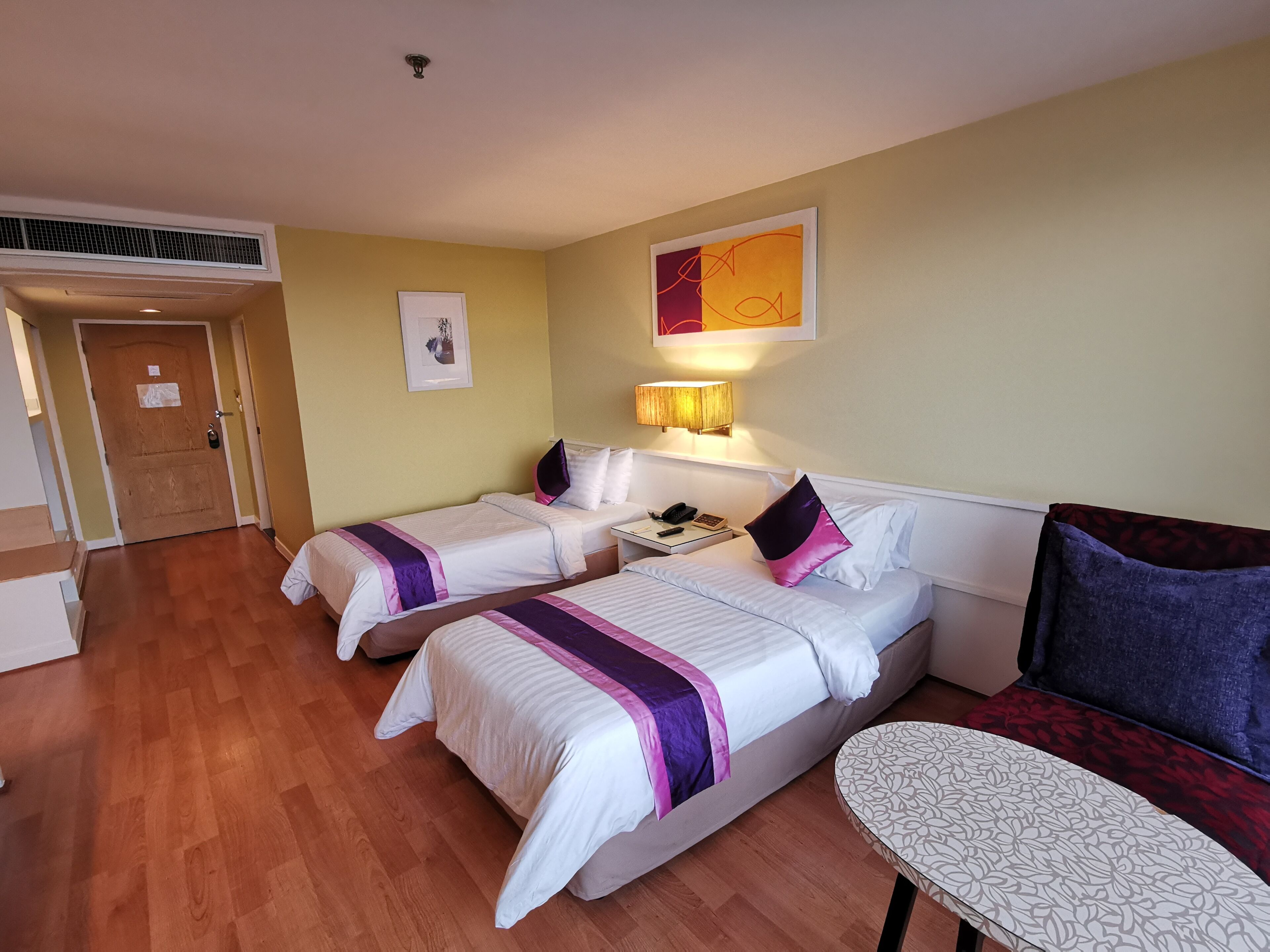 A hotel room with two beds, a sofa, a small table, and a wall-mounted lamp.