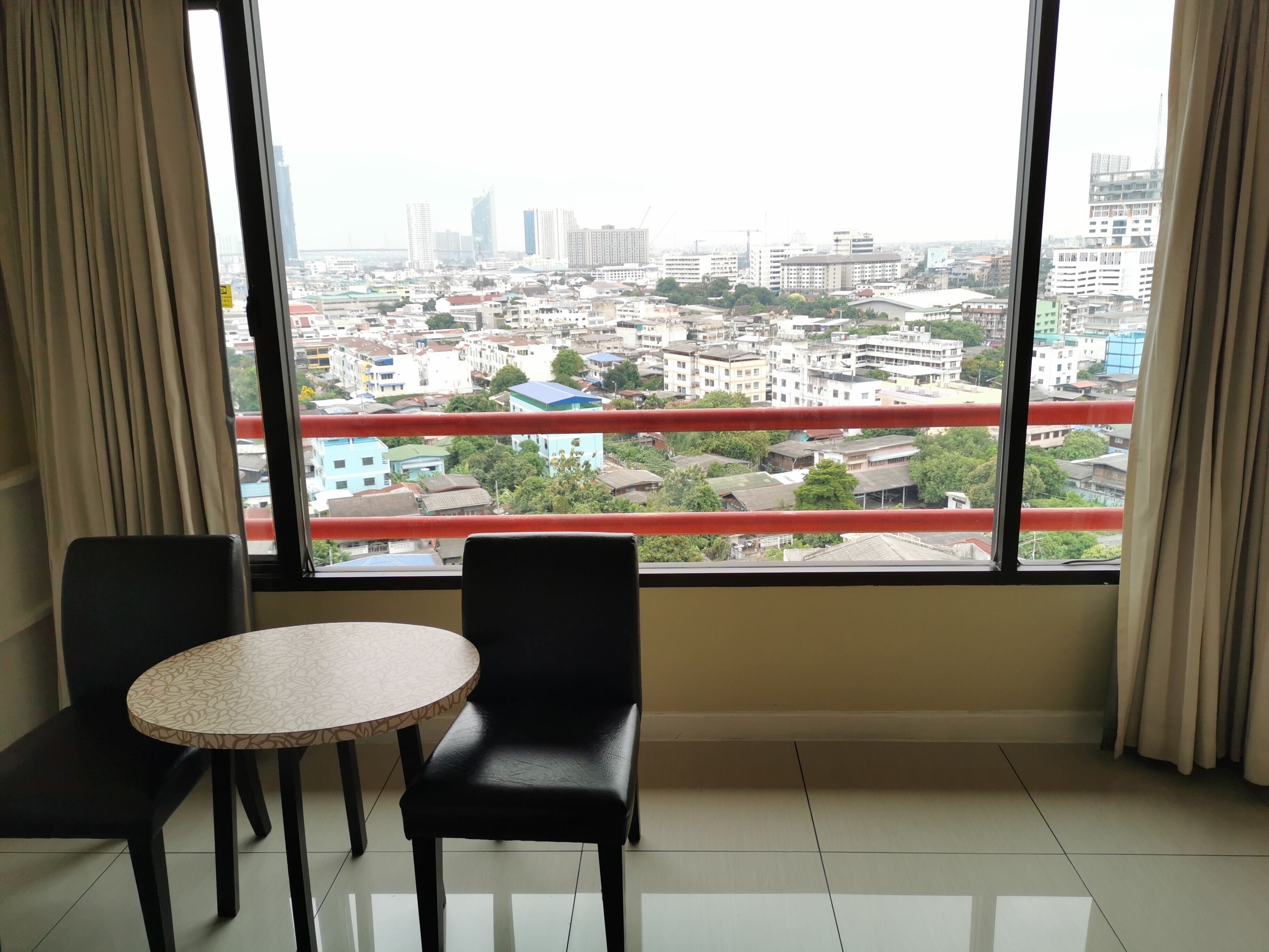 Una habitación con un ventanal amplio que ofrece vistas a la ciudad, dos sillas negras, una mesita redonda y una barandilla roja.
